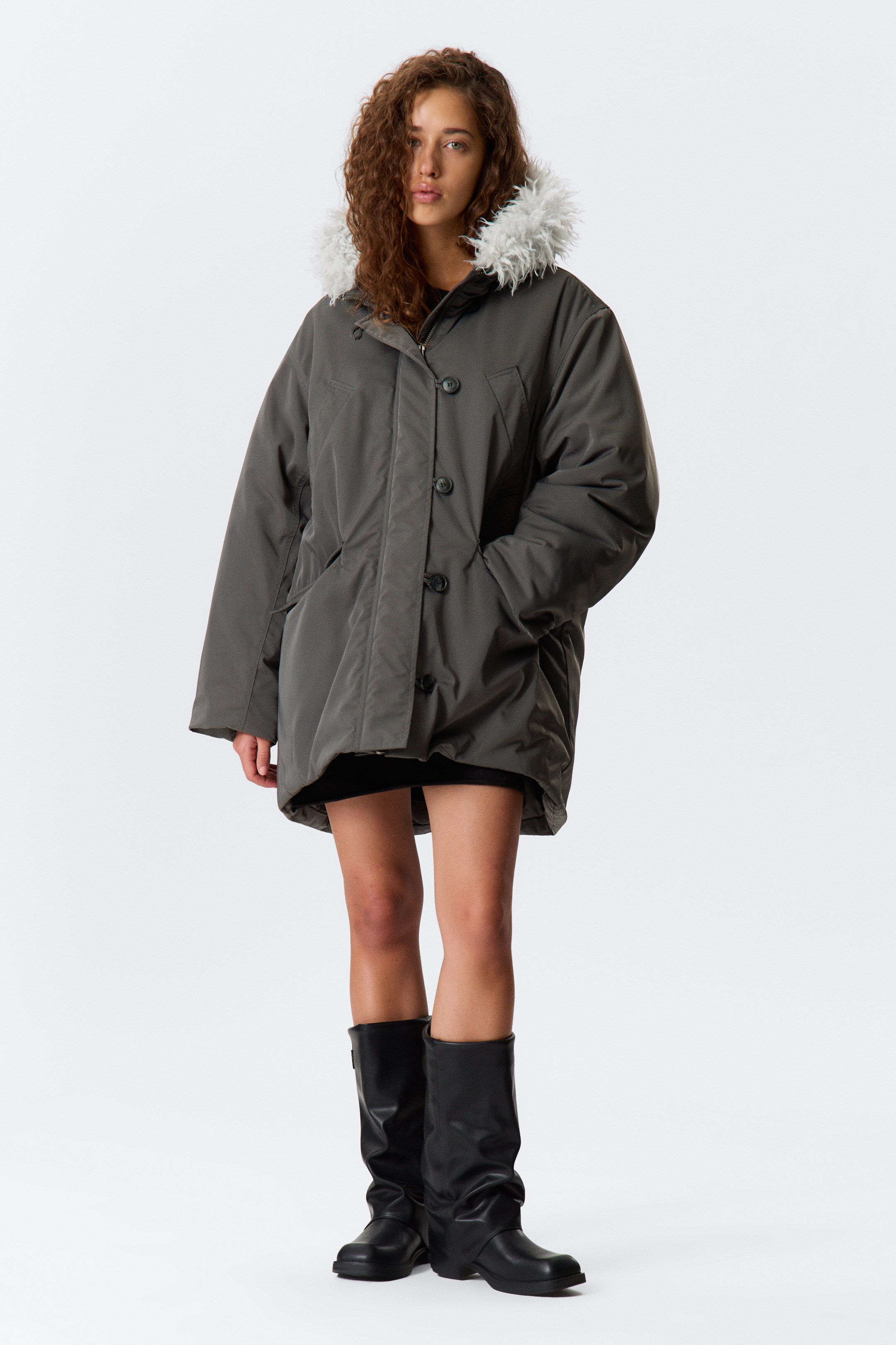 Grotere afbeelding bekijken: De houtskoolgrijze parka wordt frontaal gepresenteerd en toont een oversized silhouet, een capuchon met lichtgrijze faux fur rand, een volledige knopenlijst over een ritssluiting en twee zichtbare steekzakken aan de onderkant van de voorkant.