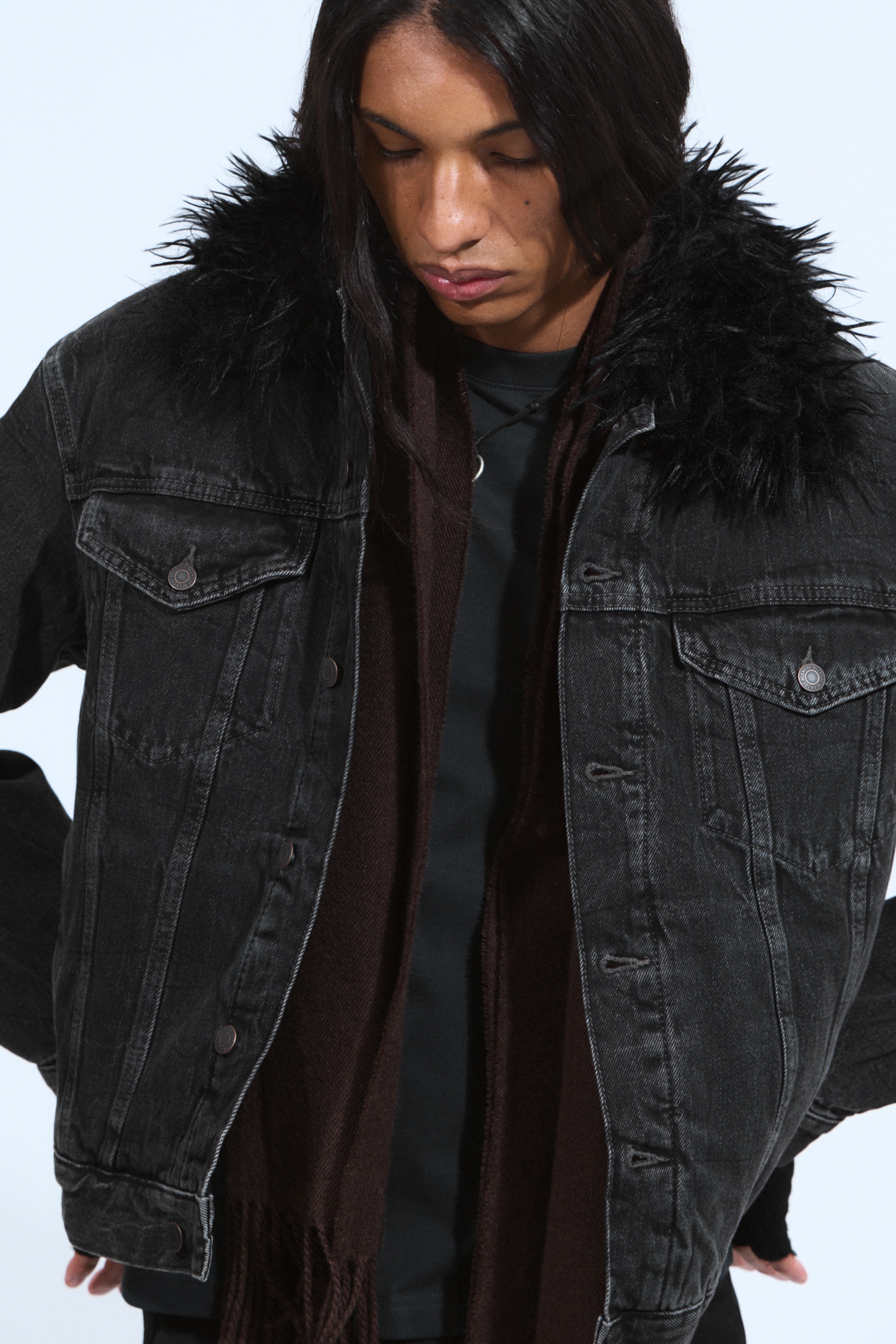View larger image: A black washed denim jacket is featuring a shaggy black faux fur collar, two buttoned chest flap pockets, and a button-front closure. It is being worn open, layered over a dark shirt and a brown fringed scarf.
