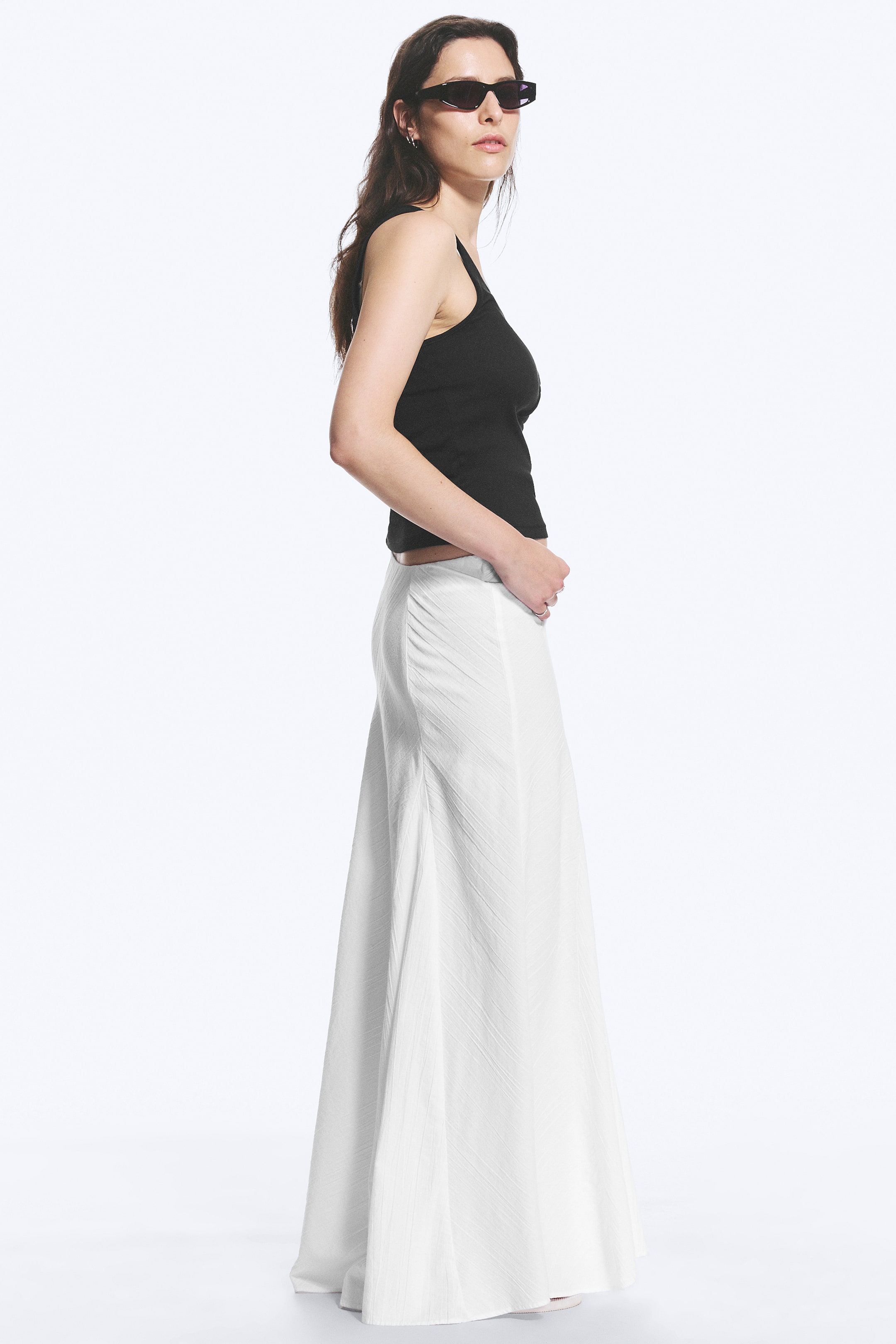 Agrandir l'image: Une jupe longue blanche plissée, avec une silhouette en ligne A et un détail froncé à la hanche, est portée par le mannequin. La main du mannequin repose sur la ceinture de la jupe.