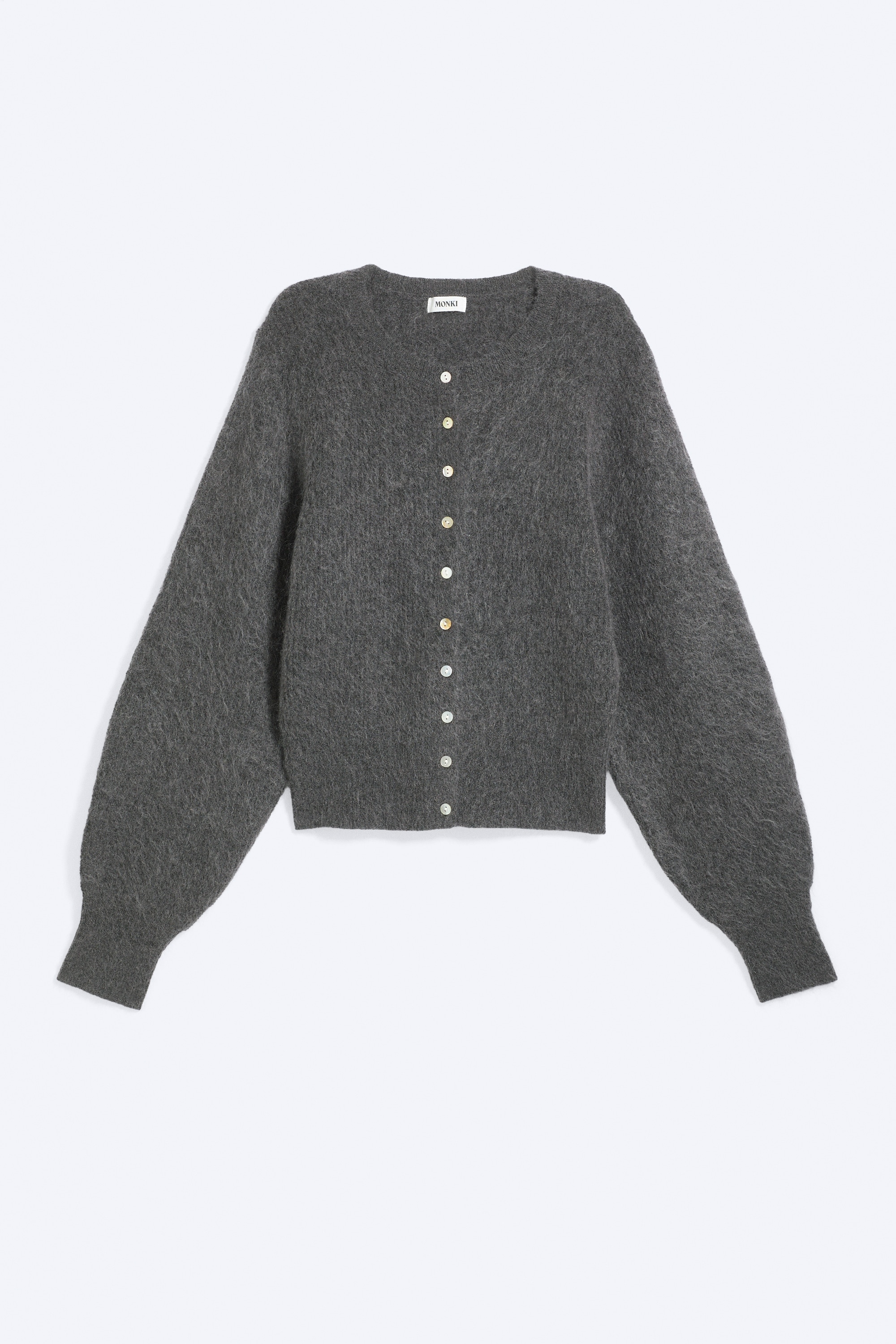 Eine graue, gebÃ¼rstete Strickjacke aus Mohair-Mischung wird vom Model Ã¼ber einem petrolfarbenen Langarmtop und braunen Hosen getragen. Das Model richtet die Vorderseite der teilweise geknÃ¶pften Strickjacke und betont damit ihren runden Ausschnitt, die kleinen weissen KnÃ¶pfe sowie die langen Ãrmel mit RippbÃ¼ndchen.