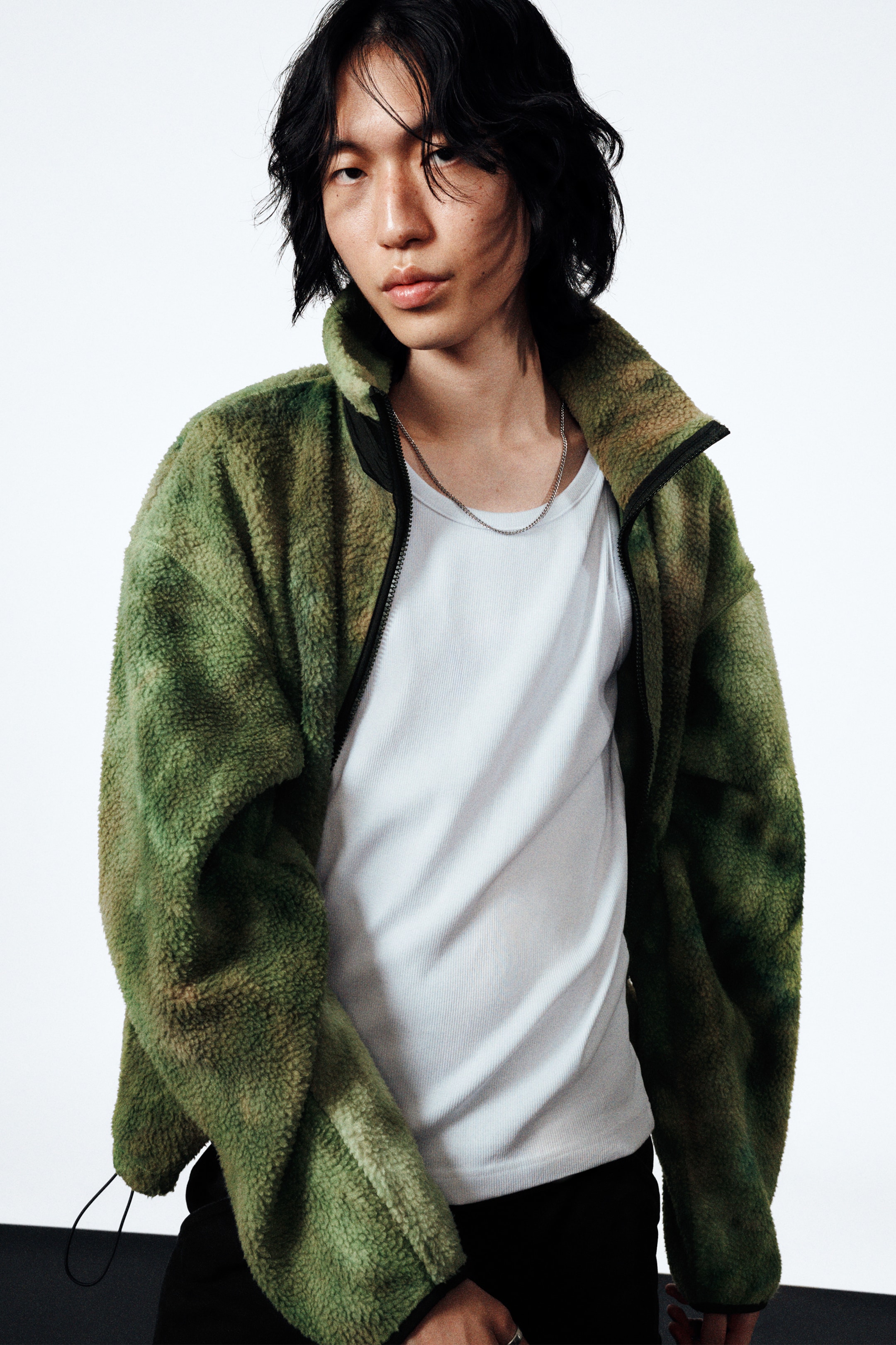 Grösseres Bild ansehen: Eine grün-braun gesprenkelte Fleecejacke mit flauschiger Textur, Stehkragen und offenem schwarzem Reissverschluss wird getragen, wobei ein weisses geripptes Tanktop darunter sichtbar wird. Ein schwarzer Kordelzug ist am Saum der Jacke sichtbar.