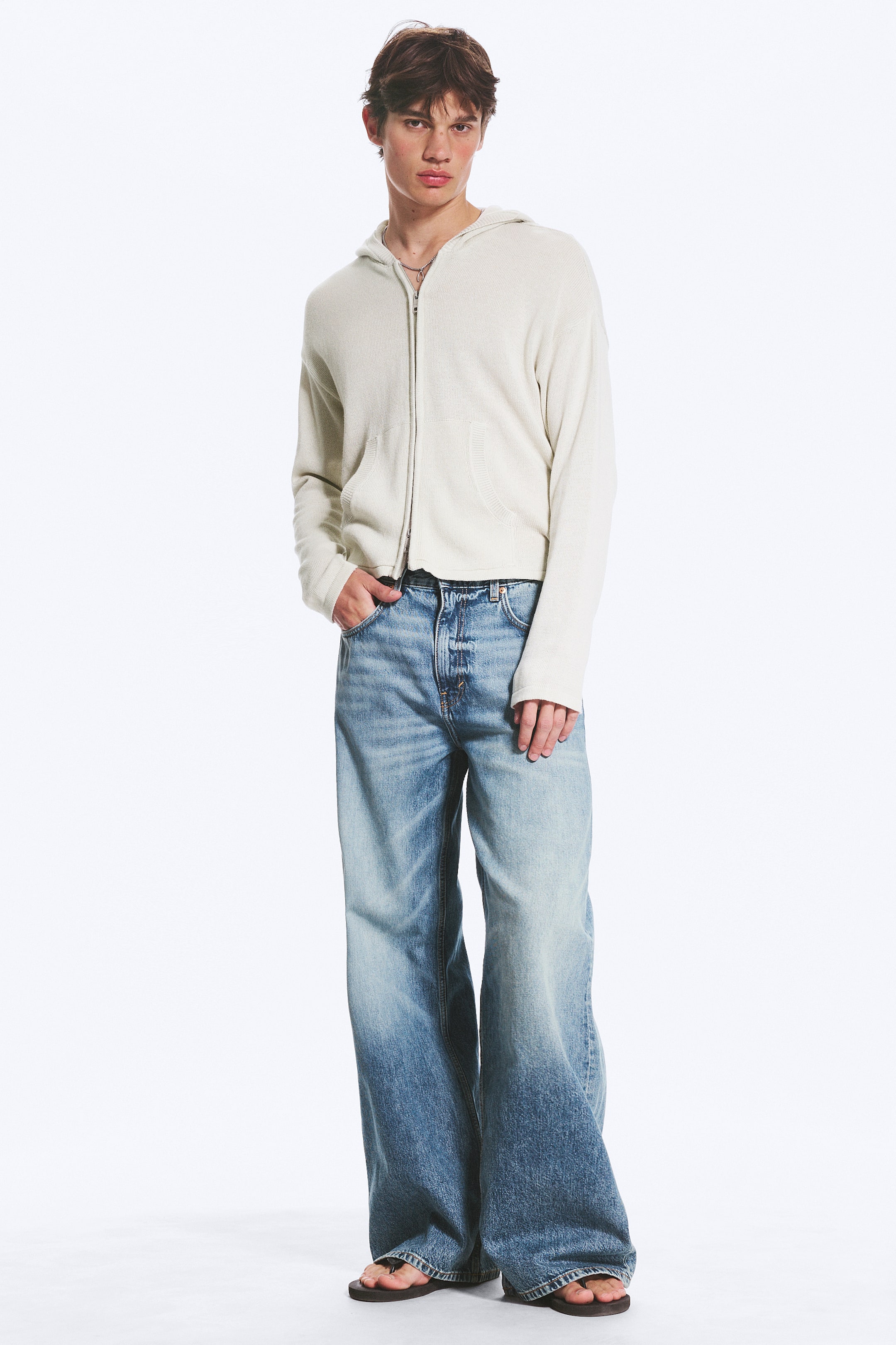Agrandir l'image: Le mannequin porte un jean à coupe ample et jambes larges avec une taille mi-haute, glissant une main dans une poche avant. Le denim rigide présente un dégradé bleu clair s'estompant vers un bleu plus foncé, avec une fermeture à boutons visible à la taille.