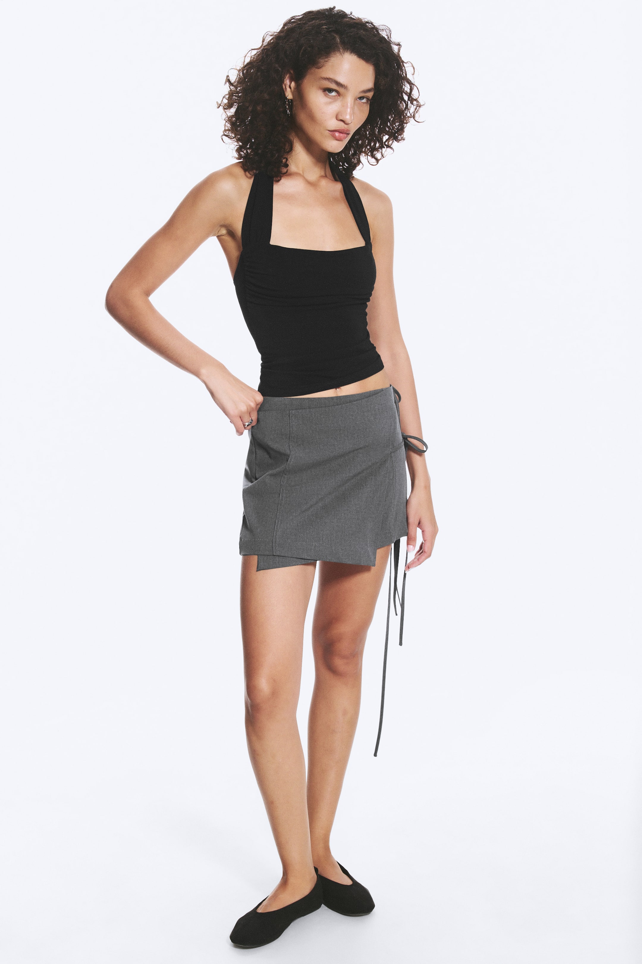 View larger image: A grey mini skirt featuring an asymmetrical, wrap-style design with overlapping panels, long black ties hanging from the left side, and visible seams detailing the front.