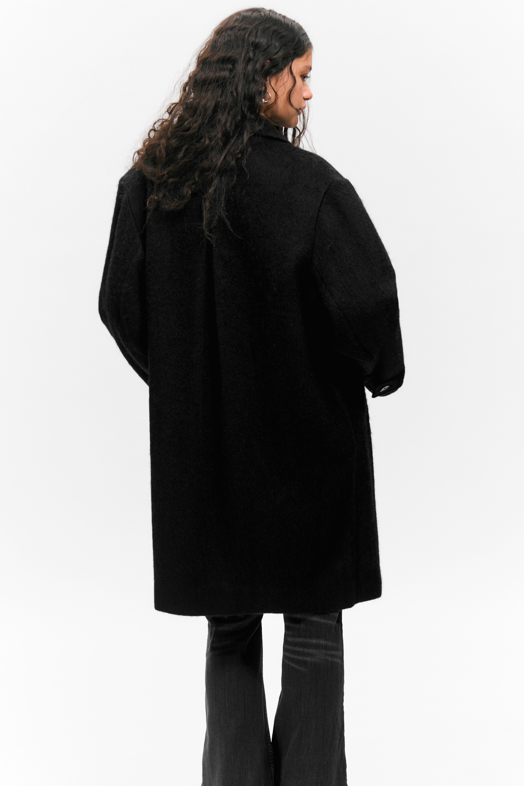 Visualizza immagine più grande: Un cappotto nero dalla texture pelosa viene mostrato dal retro, caratterizzato da una vestibilità ampia e oversize con maniche lunghe. L'orlo del cappotto cade sopra le ginocchia, coprendo parzialmente pantaloni scuri.