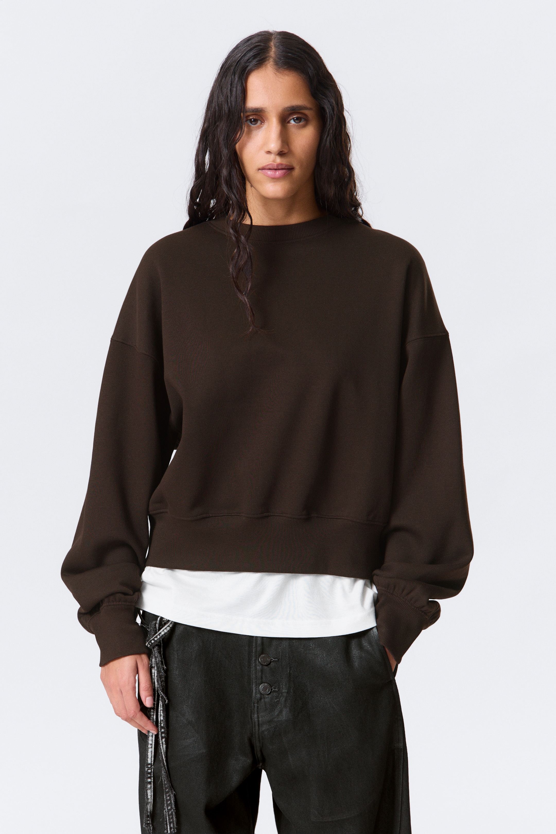Agrandir l'image: Un sweat-shirt brun foncé est vu de face, avec un col rond et des manches longues avec des poignets côtelés. Son ourlet côtelé définit une longueur raccourcie, et un t-shirt blanc est visible en dessous.
