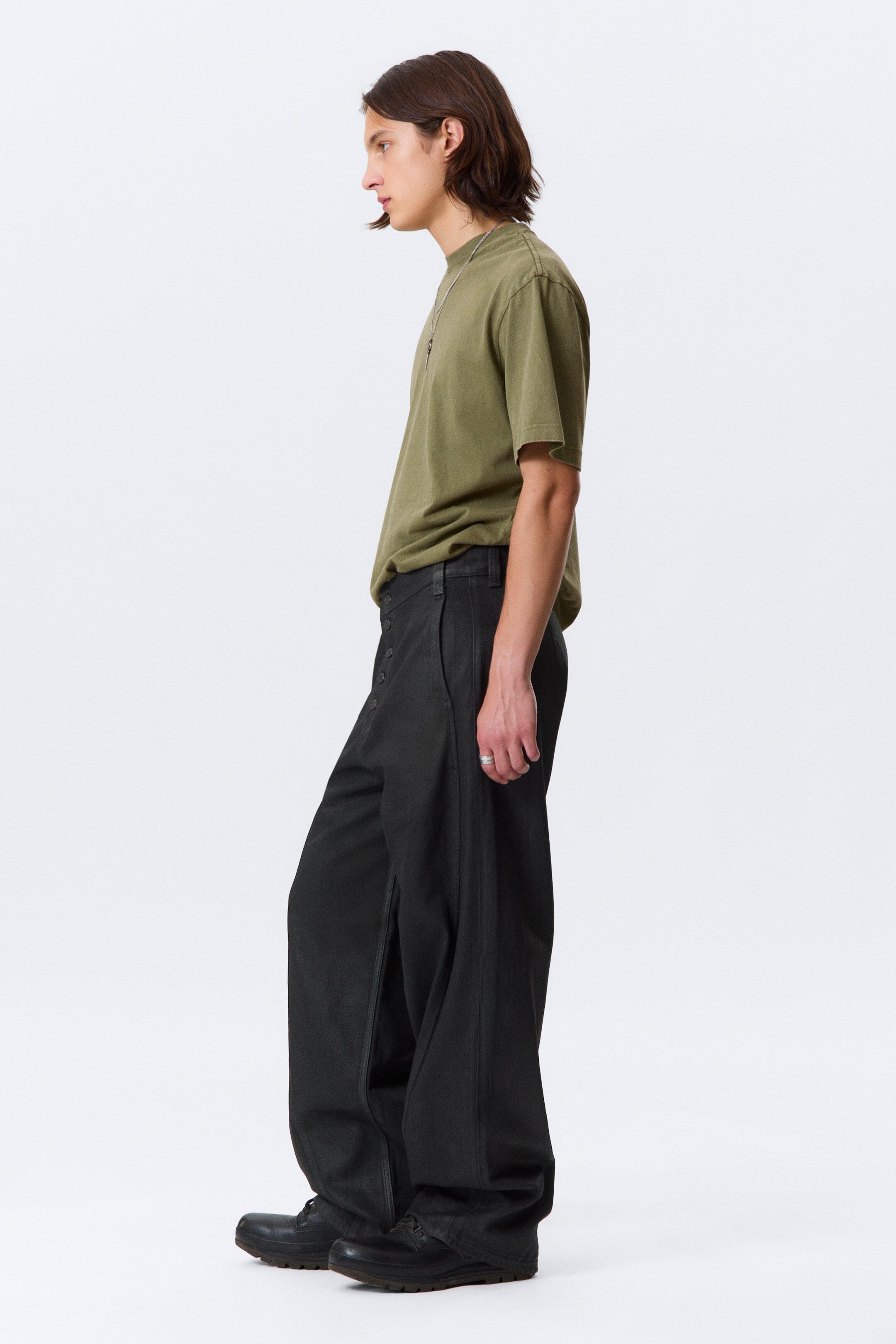 View larger image: Dark grey, wide-leg trousers are being worn by a person in a side profile, showing their loose fit and length pooling over black shoes. The trousers feature an exposed button fly closure on the side. An olive green t-shirt is being tucked into the trousers.