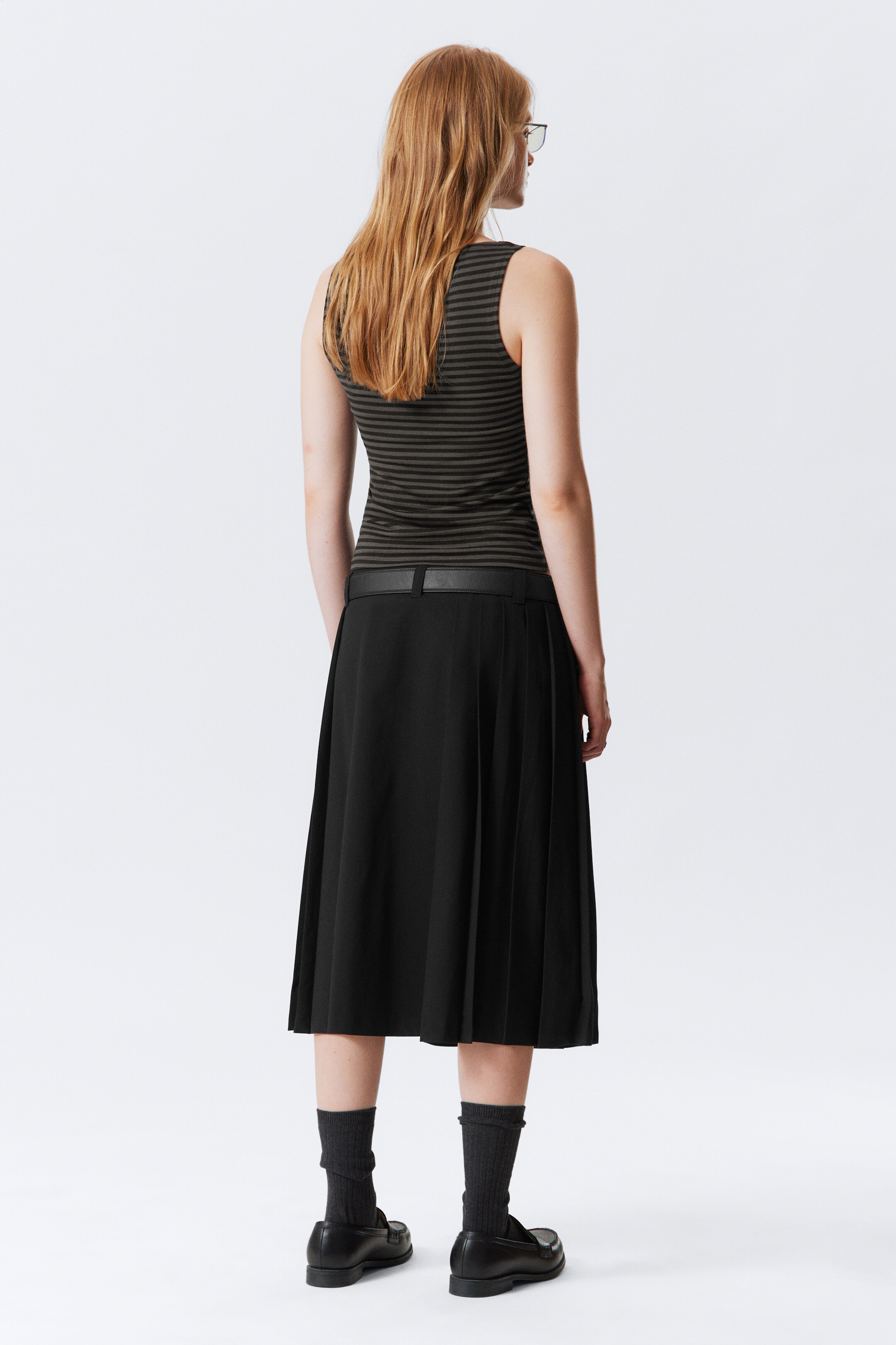 Ver imagen más grande: Se lleva una camiseta sin mangas con rayas horizontales en verde oliva y gris oscuro, vista desde la parte trasera. Está combinada con una falda midi plisada negra, ajustada en la cintura por un cinturón negro delgado. Se llevan calcetines de canalé gris oscuro y mocasines negros.