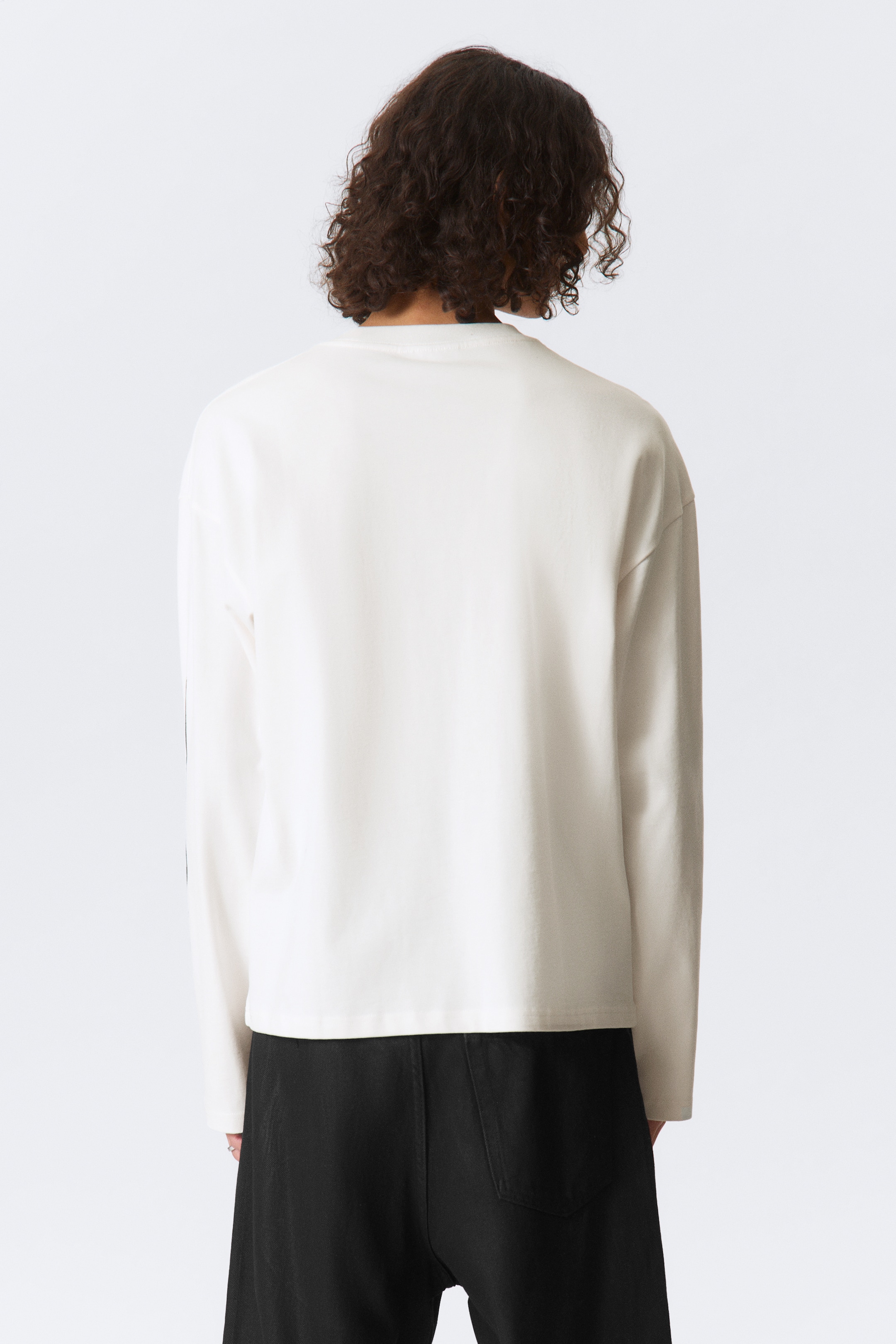 Vis større bilde: En off-white langermet topp vises fra baksiden. Den har en rund hals og en avslappet, rett passform, med lange ermer som ender i enkle mansjetter og en rett kant.