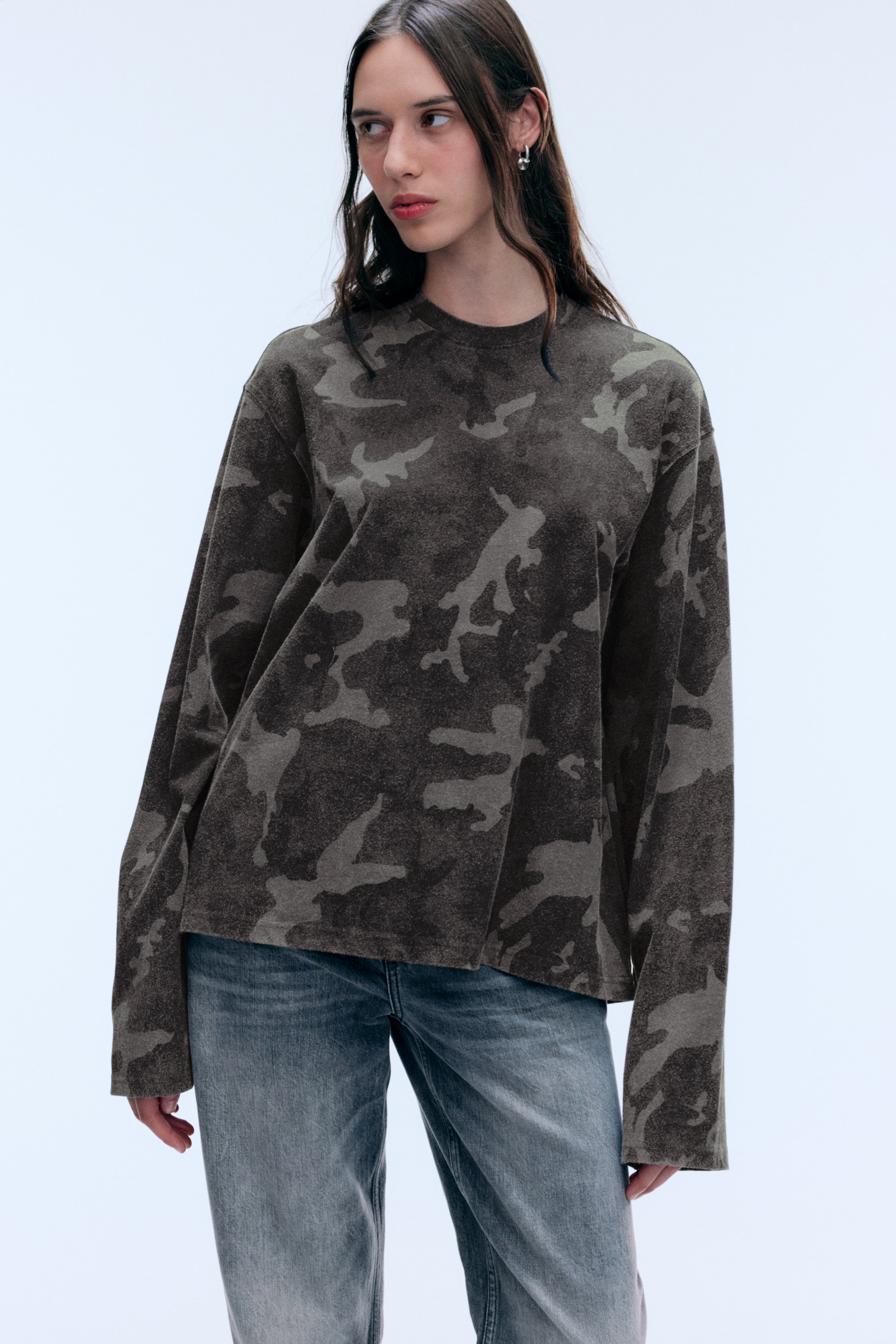 Ver imagen más grande: La modelo lleva puesta una prenda de manga larga estampada con un patrón de camuflaje oscuro. La prenda de corte relajado tiene cuello redondo y mangas largas, y se combina con unos vaqueros denim grises.