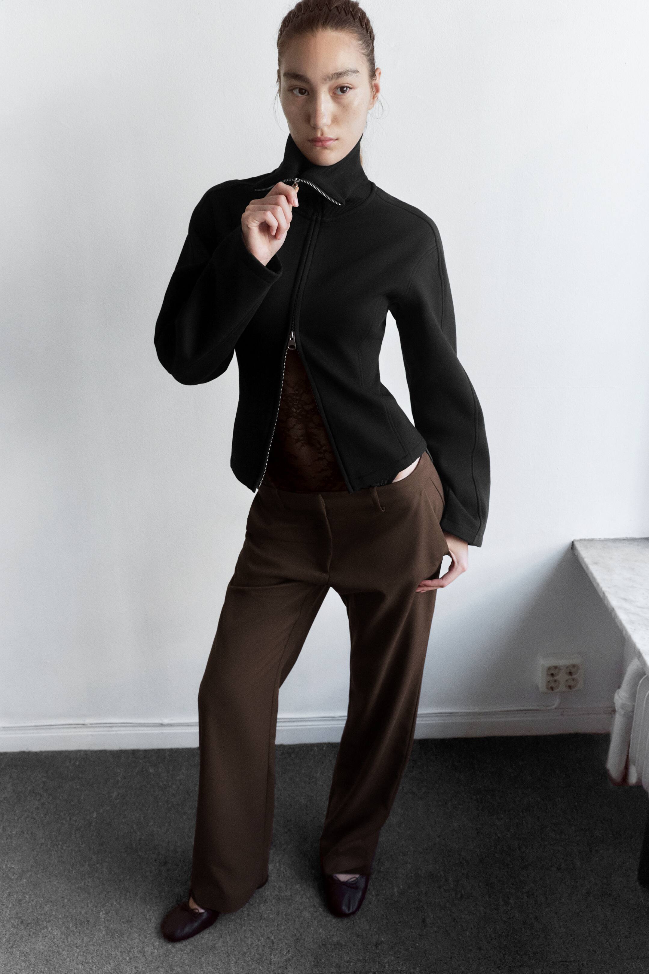 Visualizza immagine più grande: Una felpa nera slim fit con colletto alto, cuciture a pannelli e zip a doppio cursore è indossata dalla modella, che sta regolando il tiralampo superiore. La felpa è abbinata con la zip inferiore parzialmente aperta per rivelare un sottostrato in pizzo scuro, indossata con pantaloni marroni a gamba larga.
