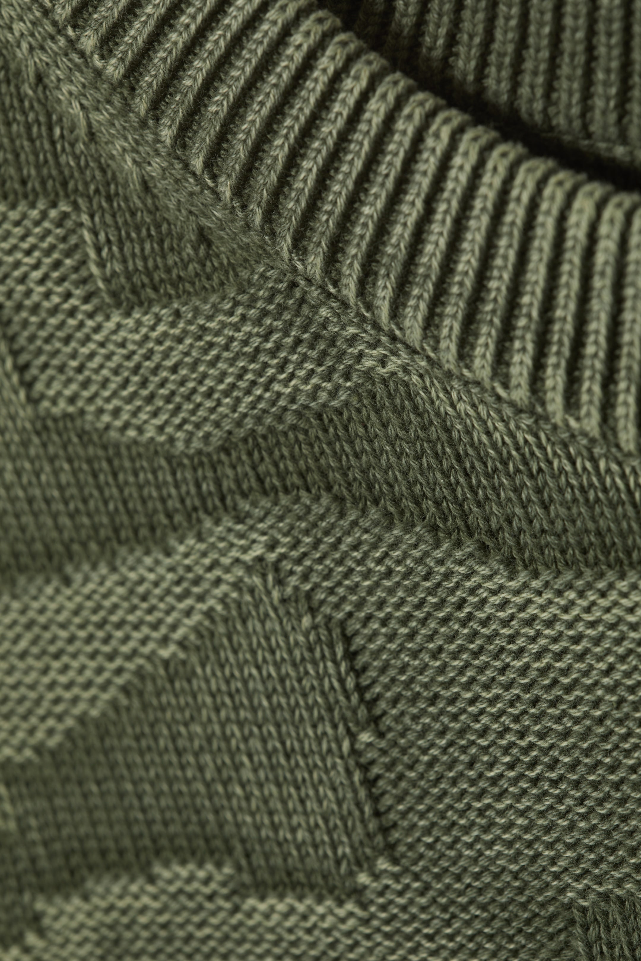 Agrandir l'image: Un gros plan d'un tissu tricoté vert olive présente une large bordure côtelée en haut, avec le corps principal affichant un motif texturé répétitif de dessins en chevrons surélevés.