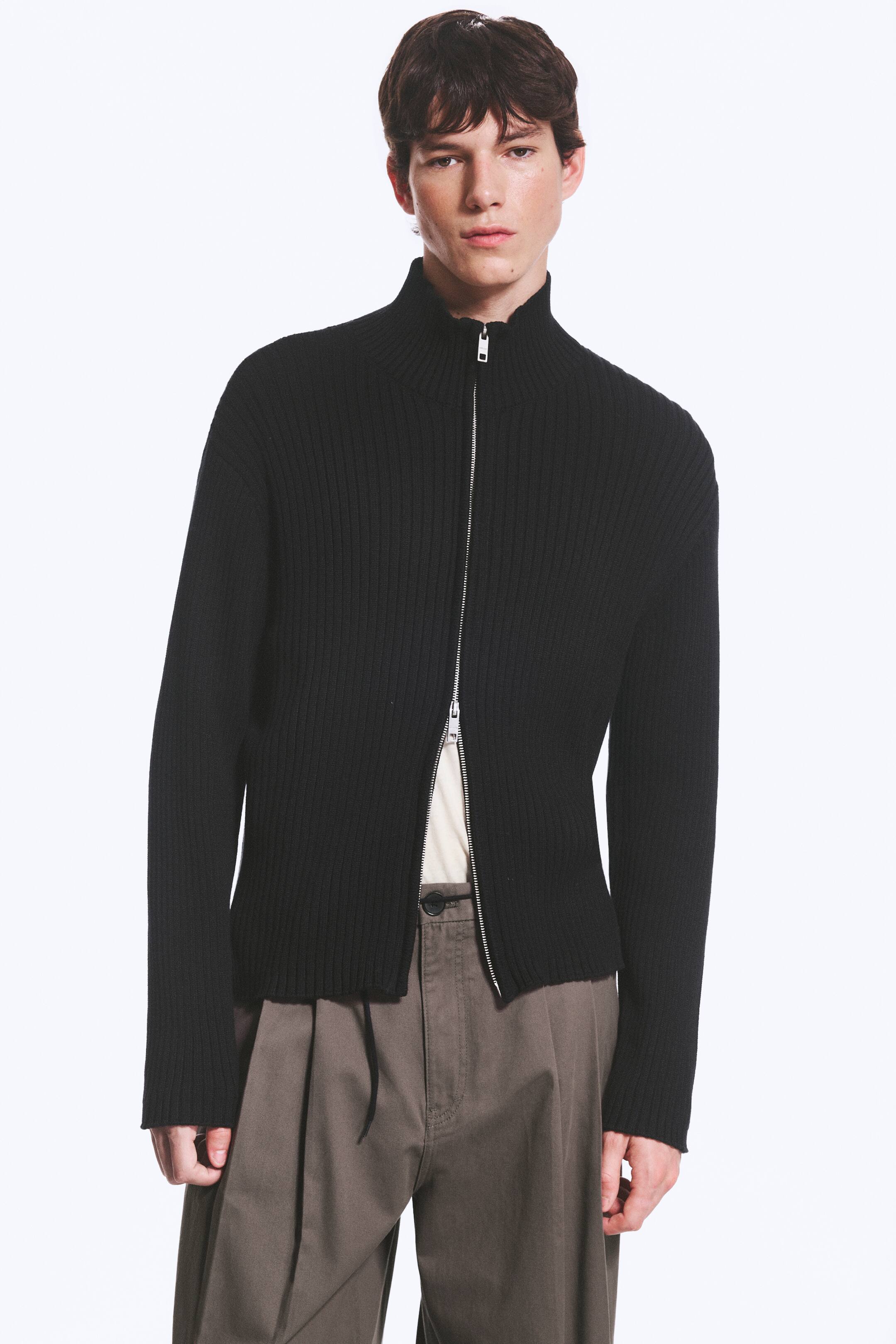 Visualizza immagine più grande: Un cardigan nero a vestibilità rilassata, caratterizzato da una texture a coste e un collo alto, è indossato dalla modella. La chiusura con zip a doppio cursore è parzialmente aperta da entrambe le estremità, rivelando una maglia di colore chiaro sotto.
