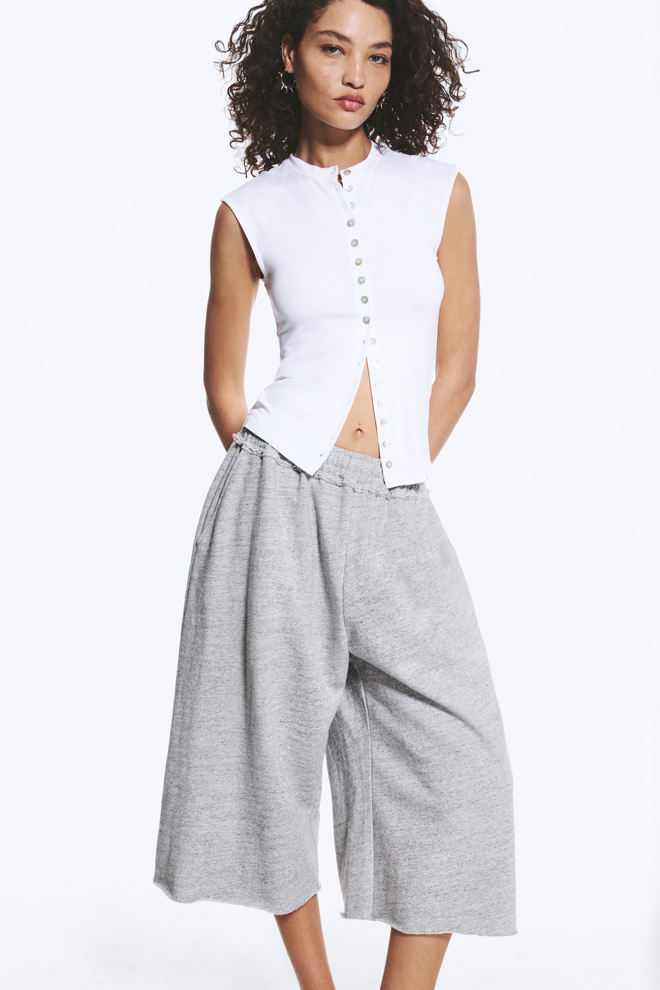 Se større billede: En hvid ærmeløs ribstrikket top med båndkrave og fuld knaplukning præsenteres sammen med grå, brede, cropped sweatpants. Toppen er delvist opknappet og afslører taljen, mens bukserne fremviser en elastisk talje og rå kanter.