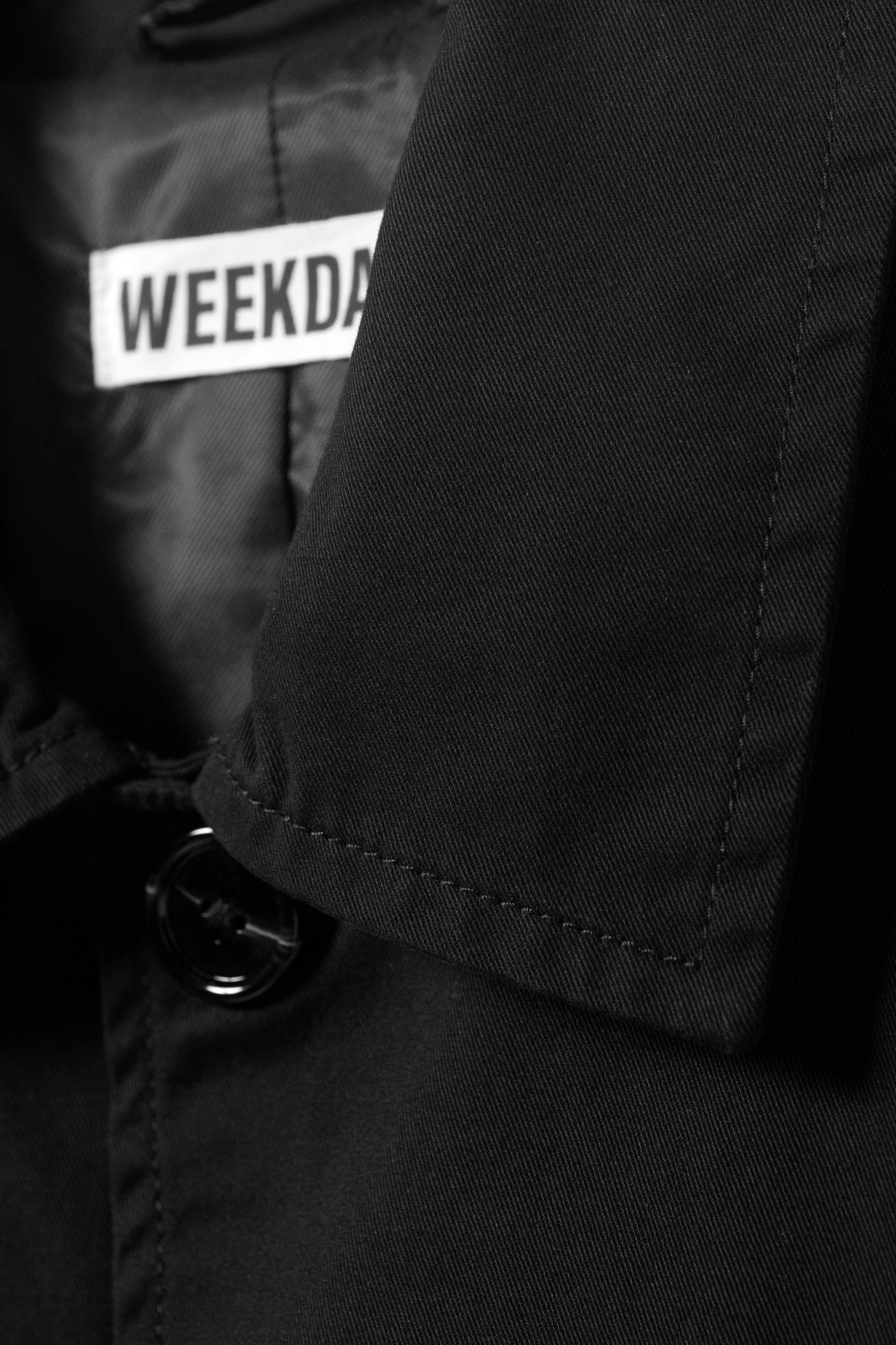 Agrandir l'image: Une veste noire est présentée en gros plan sur son col et sa fermeture avant. Le col présente des coutures visibles le long de son bord, et un bouton noir brillant sécurise le vêtement, avec une étiquette de marque "WEEKDA" blanche partiellement visible à l'intérieur.