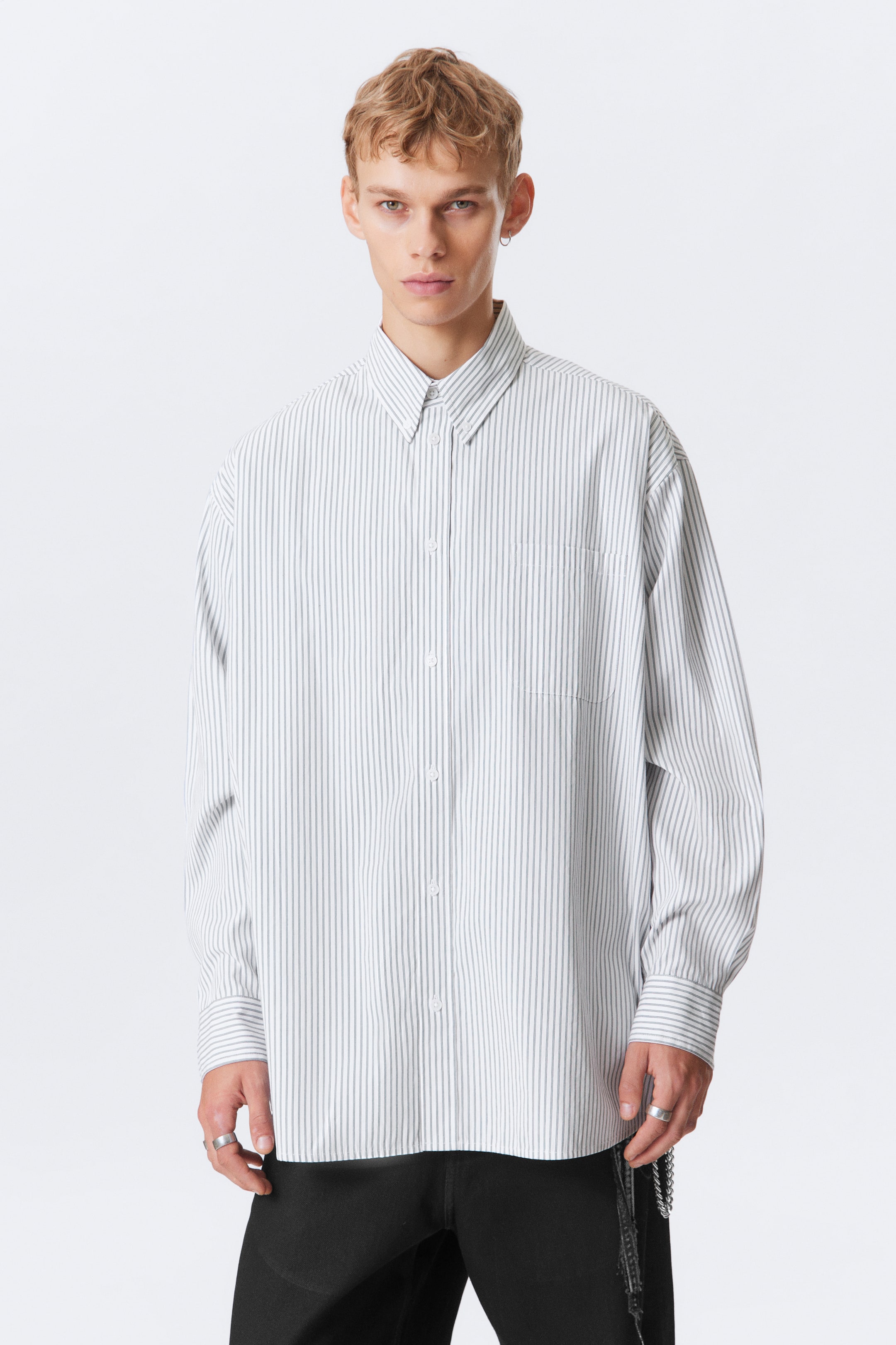 Ver imagem maior: Uma camisa de manga comprida com padrão de riscas verticais está a ser usada pelo modelo, apresentando um corte oversize, uma gola de botões, um bolso no peito, punhos com botões e uma barra curva.