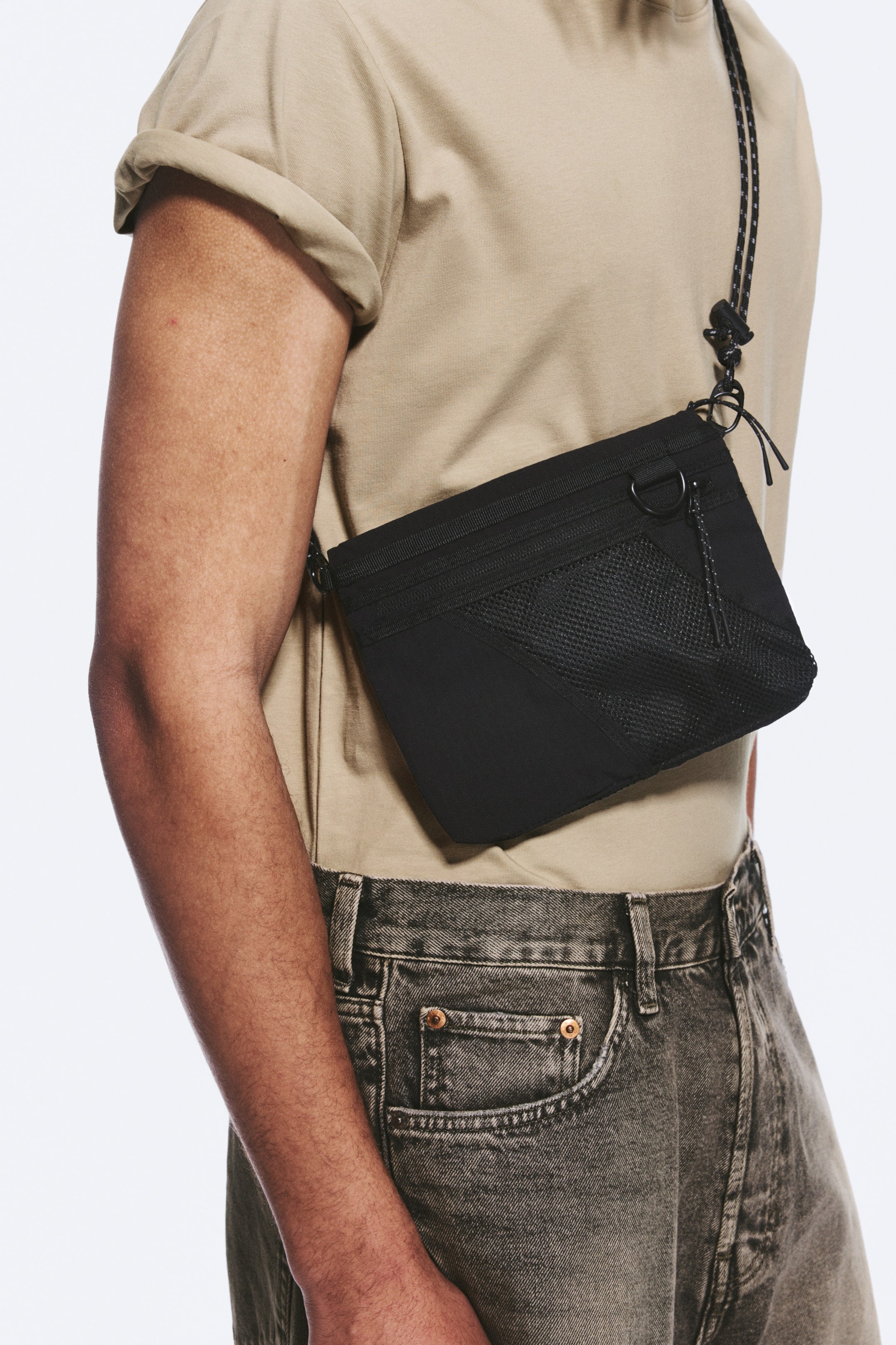 Grotere afbeelding bekijken: De zwarte crossbody tas wordt gedragen, schuin naar beneden van de linkerschouder naar de rechterheup, met een rechthoekige vorm. Hij is voorzien van een bovenste ritssluiting, een voorvak van mesh en een gevlochten koordriem die aan D-ringen op de bovenste hoeken is bevestigd.