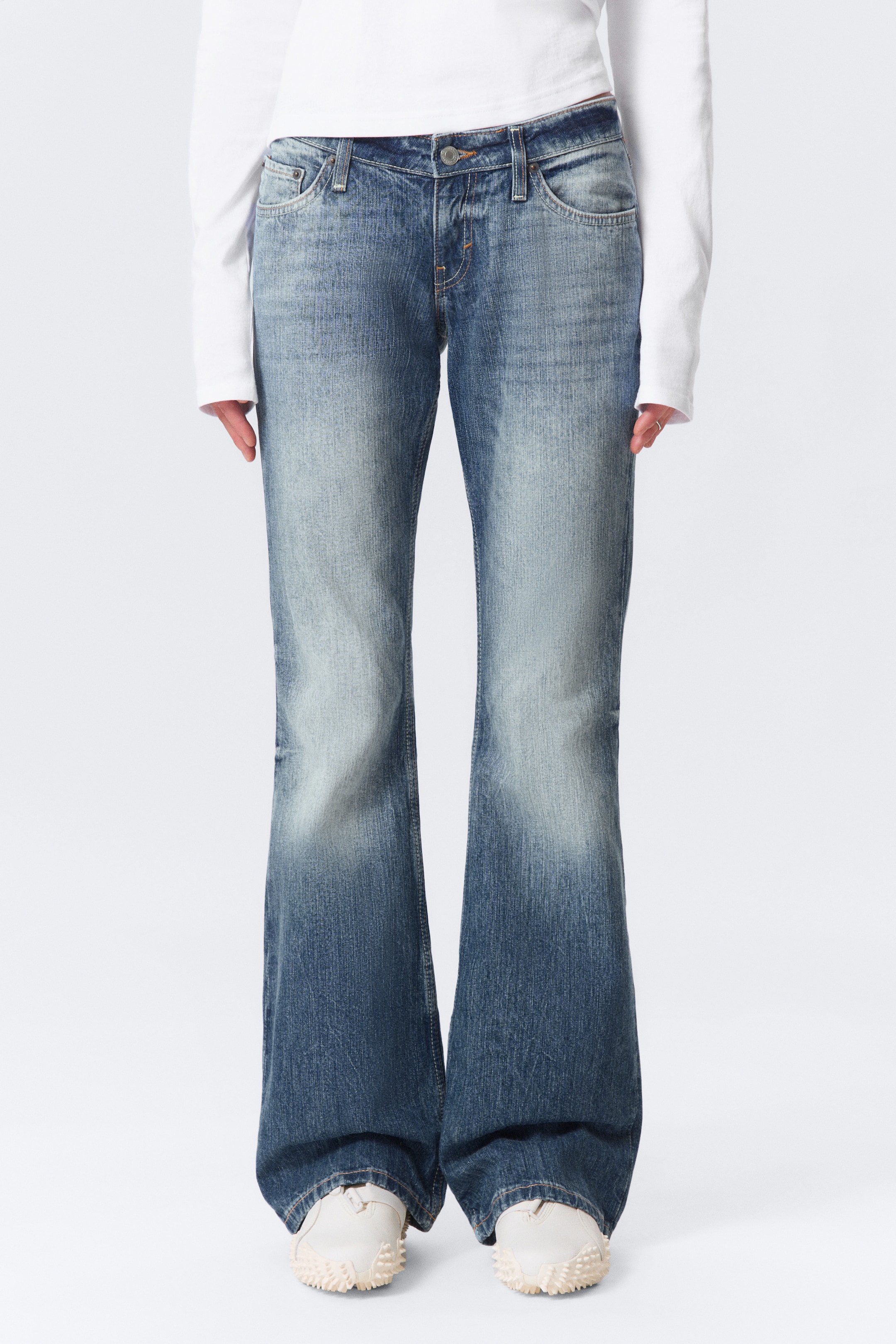 Grotere afbeelding bekijken: Low rise, slim fit, bootcut jeans in gestreepte blauwe denim worden gedragen door het model, met hun tailleband zichtbaar boven een wit langemouwen shirt. De jeans hebben een 5-pocket ontwerp, een ritssluiting en een lange lengte die witte schoenen bedekt.
