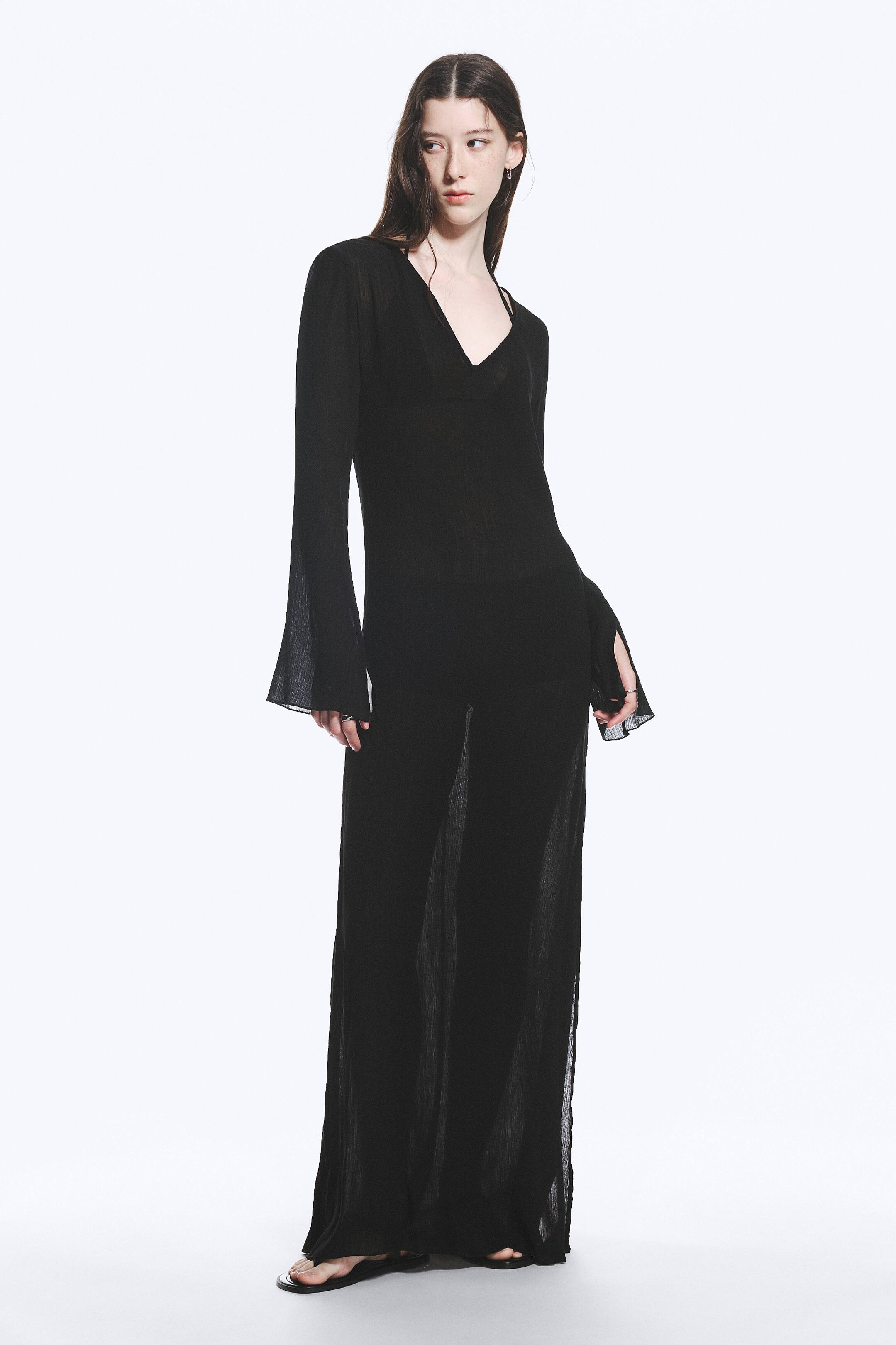 Grösseres Bild ansehen: Ein schwarzes, durchsichtiges Maxikleid ist nach vorne gerichtet und verfügt über einen V-Ausschnitt sowie lange Ärmel mit ausgestellten, geschlitzten Manschetten. Der leichte, strukturierte Stoff erzeugt eine fließende Silhouette, die durch seine transparente Verarbeitung eine innere Schicht offenbart und bis zum Boden reicht.