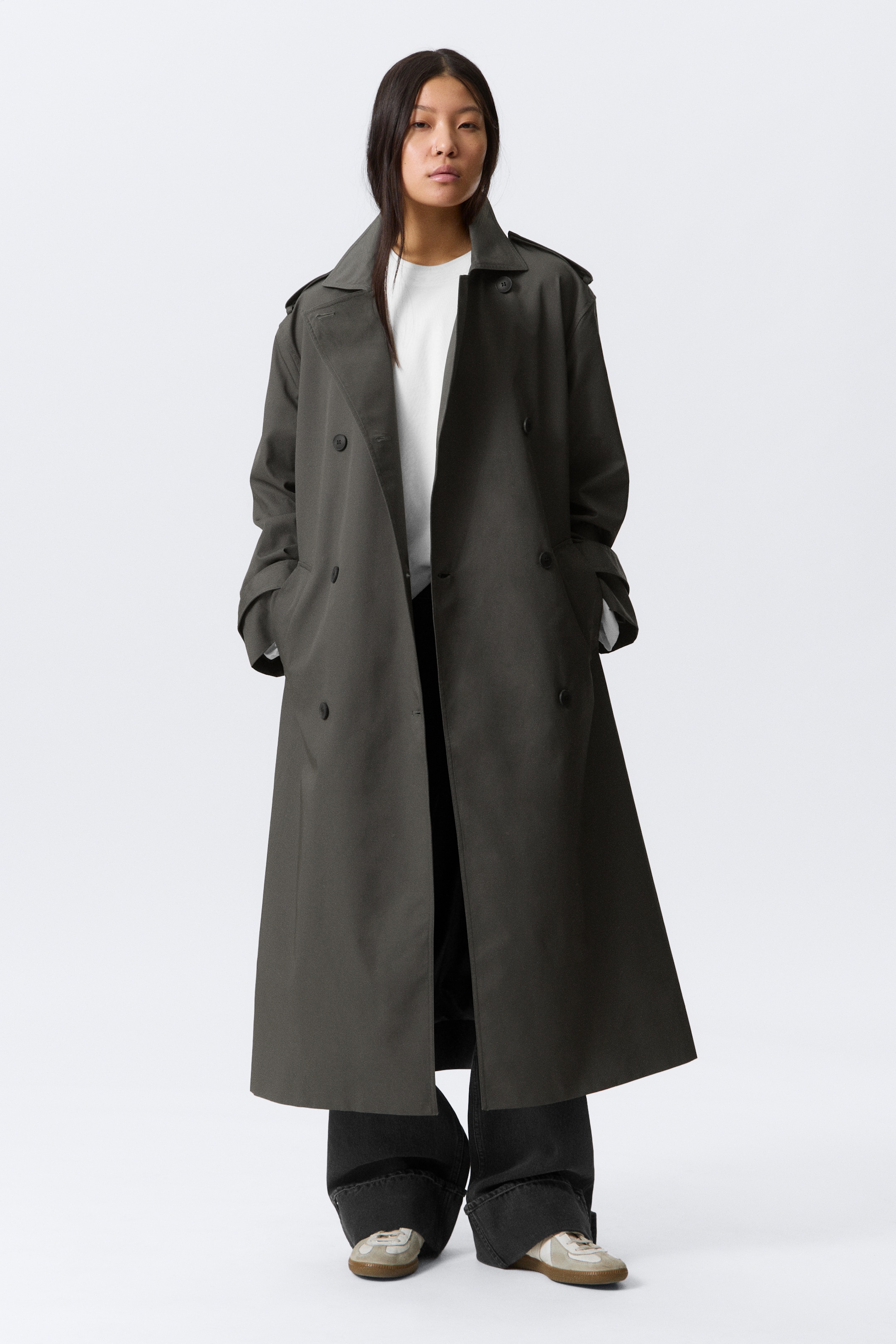 View larger image: A dark grey double-breasted trench coat features a relaxed, long silhouette with shoulder epaulets and notched lapels. It showcases side pockets with hands resting inside, and its cuffs are slightly rolled, revealing a white t-shirt worn underneath.