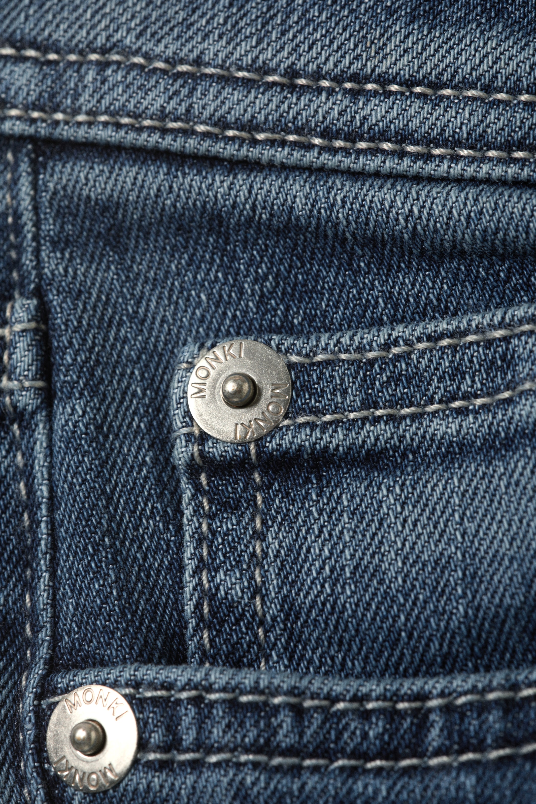 Größeres Bild ansehen: Ein blaues Denim-Kleidungsstück, von vorne gesehen, zeigt seine Köperbindung, detailliert mit weißer Steppnaht und zwei silberfarbenen Metallnieten. Die Nieten, die sich an den Taschenbereichen befinden, zeigen "MONKI" um ihre Ränder geprägt.