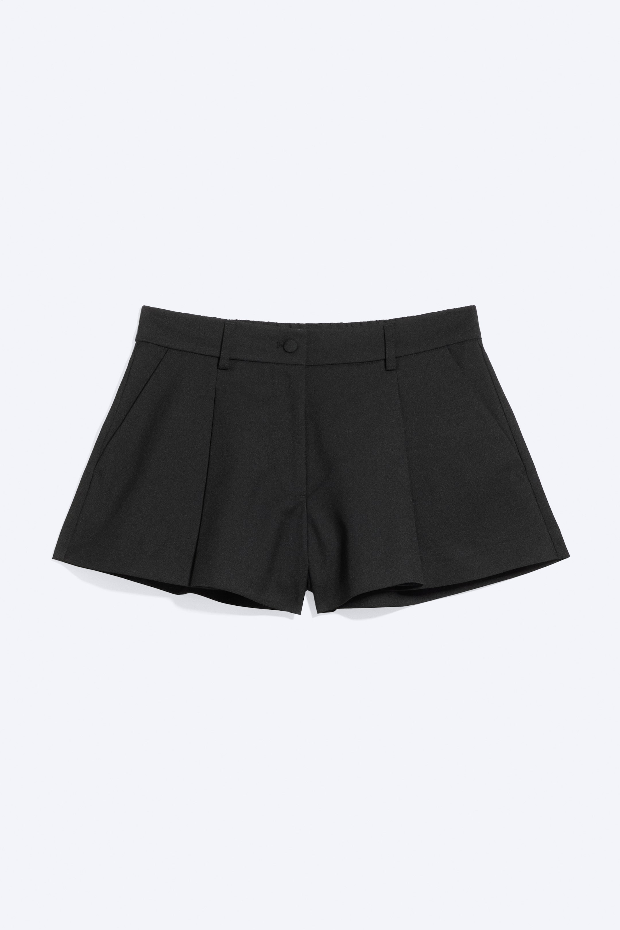 Agrandir l'image: Un short de costume noir est présenté de face, doté d'une taille mi-haute avec fermeture par bouton et passants pour ceinture. Il est conçu avec des plis à l'avant et des poches latérales en biais.