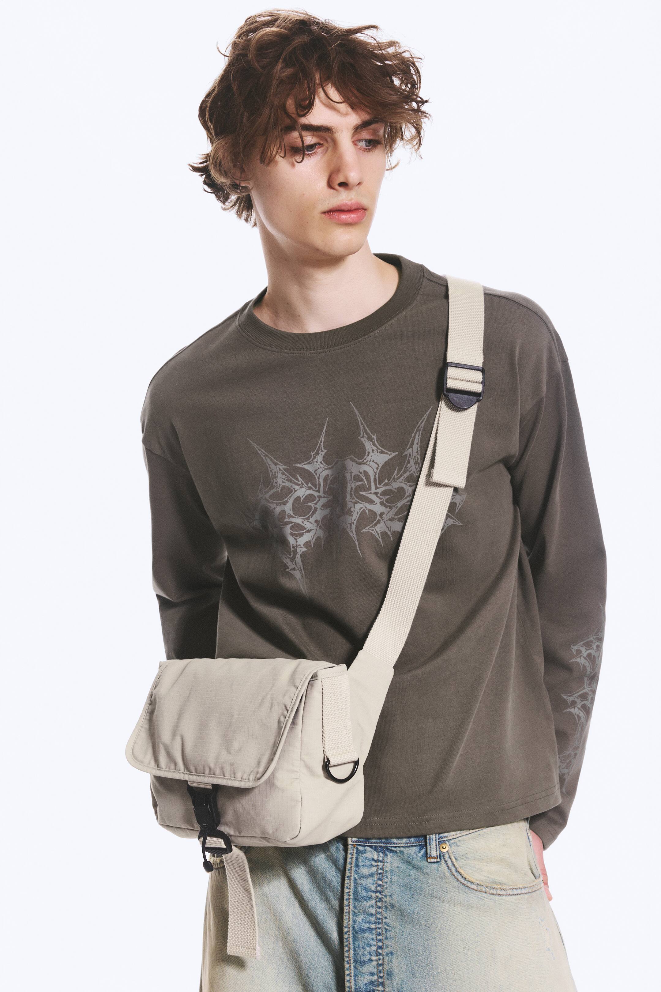 Grotere afbeelding bekijken: Een licht beige geweven stoffen crossbody tas wordt over het lichaam gedragen en rust op de rechterheup van de drager. De tas heeft een rechthoekig hoofdvak met een voorflap die wordt vastgezet door een zwarte kunststof gesp, en een brede verstelbare stoffen riem.