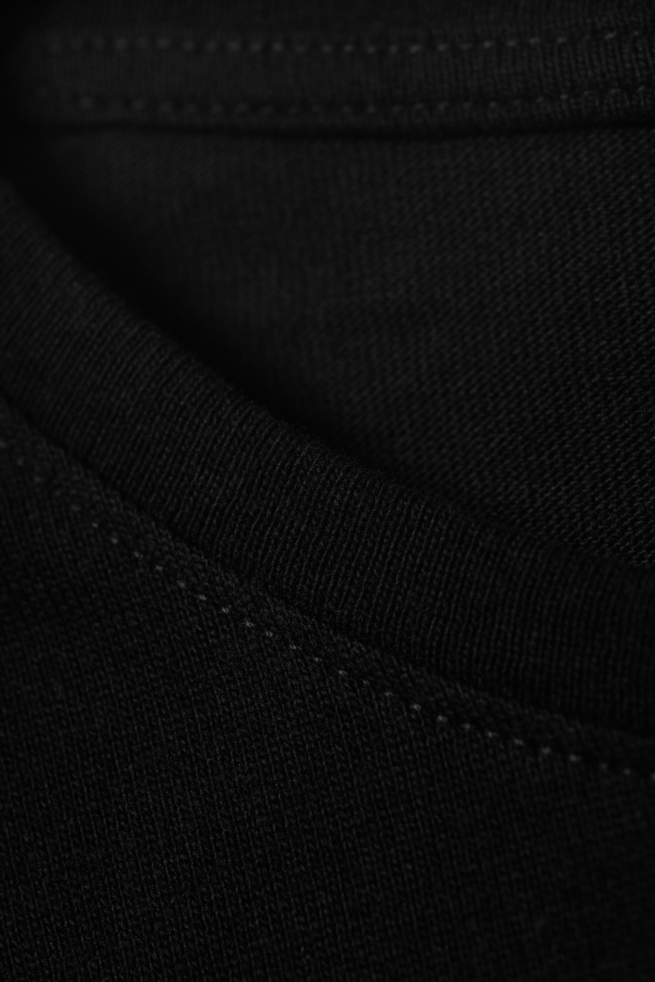 Grösseres Bild ansehen: Ein schwarzes Jersey-Strickkleidungsstück zeigt seine weiche Stoffstruktur mit subtilen Falten. Eine doppelte Reihe von hellfarbigen Steppnähten verläuft entlang einer sichtbaren Naht.