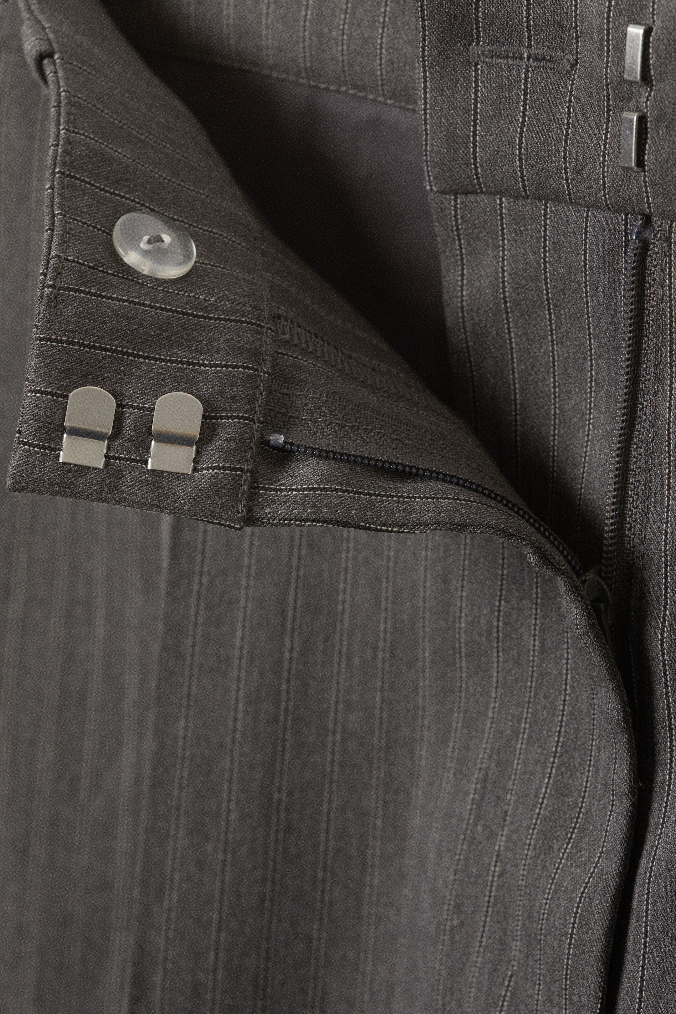 Ingrandisci l'immagine: Un paio di pantaloni a righe sottili grigio scuro viene mostrato, con un'attenzione particolare alla cerniera parzialmente aperta. Due chiusure a gancio e occhiello in metallo color argento fissano la cintura, e un bottone traslucido è visibile sulla cintura interna.