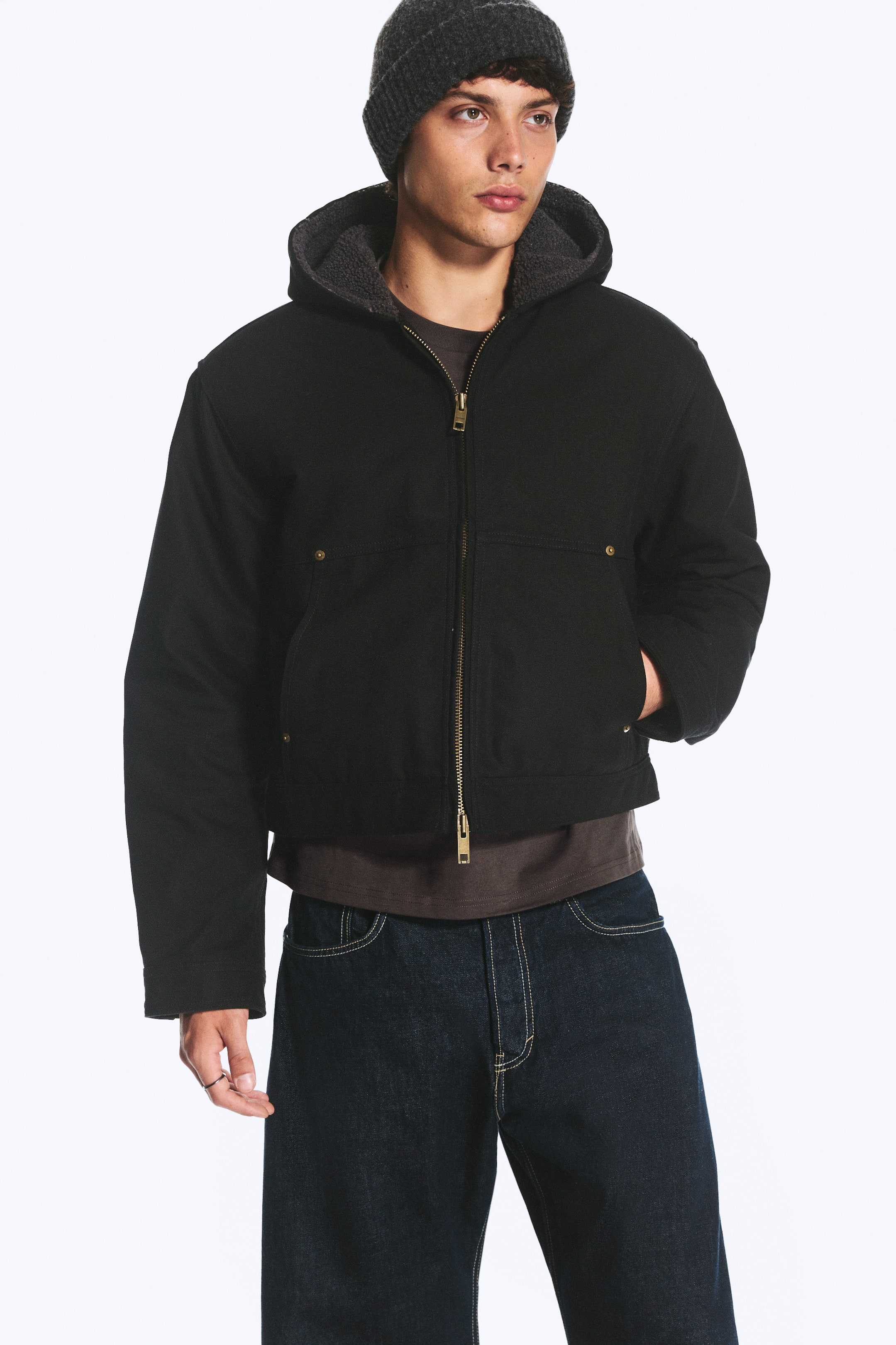 View larger image: A black hooded utility jacket featuring a gold-toned front zip, rivet-detailed pockets, and a grey sherpa-lined hood is being worn by the model. The jacket is layered over a dark T-shirt and dark denim jeans, with the model placing one hand in a side pocket.