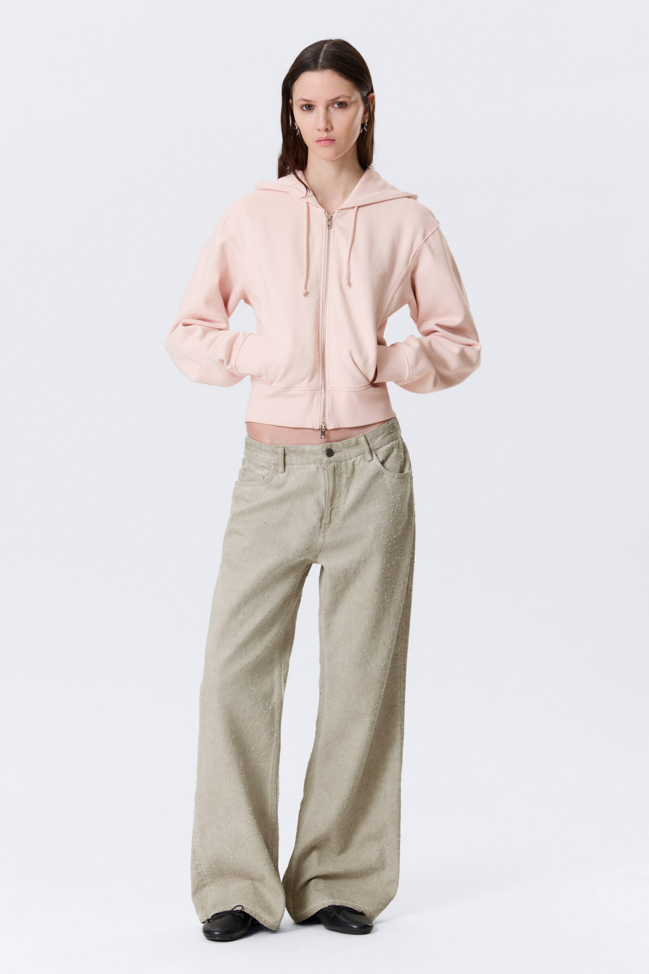 Grotere afbeelding bekijken: Licht beige broek met wijde pijpen vertoont een nubby textuur, voorzakken en een knoopsluiting aan de taille, met de tailleband zichtbaar onder een cropped top, wat een ontspannen, full-length pasvorm presenteert.