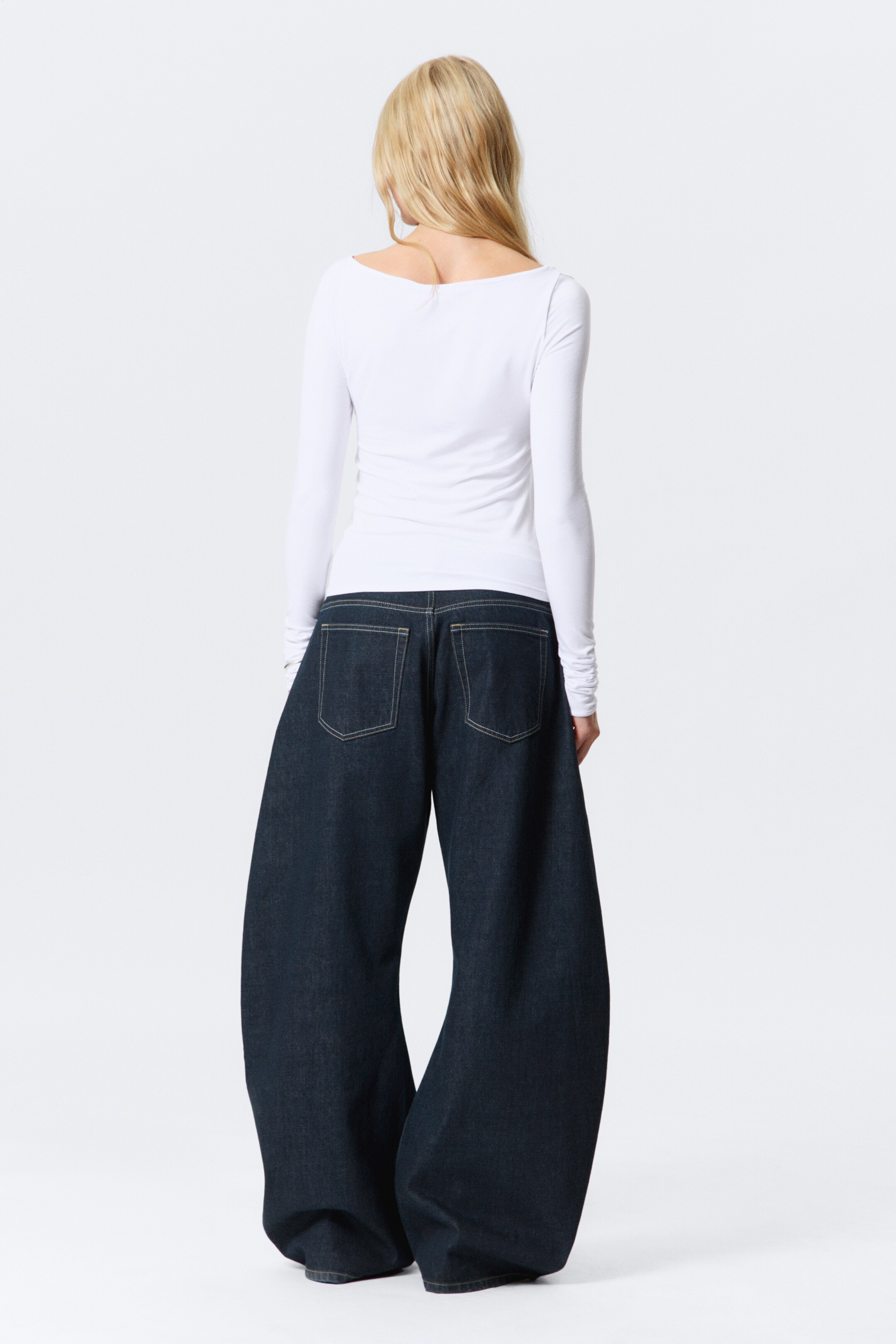 Grösseres Bild ansehen: Ein weisses Langarmoberteil und dunkel gewaschene Jeans werden von hinten gezeigt. Das enganliegende weisse Oberteil hat einen breiten Bootsausschnitt, während die dunkel gewaschenen Jeans eine sehr weite, voluminöse Silhouette mit kontrastierenden hellen Nähten und zwei aufgesetzten Gesässtaschen präsentieren.