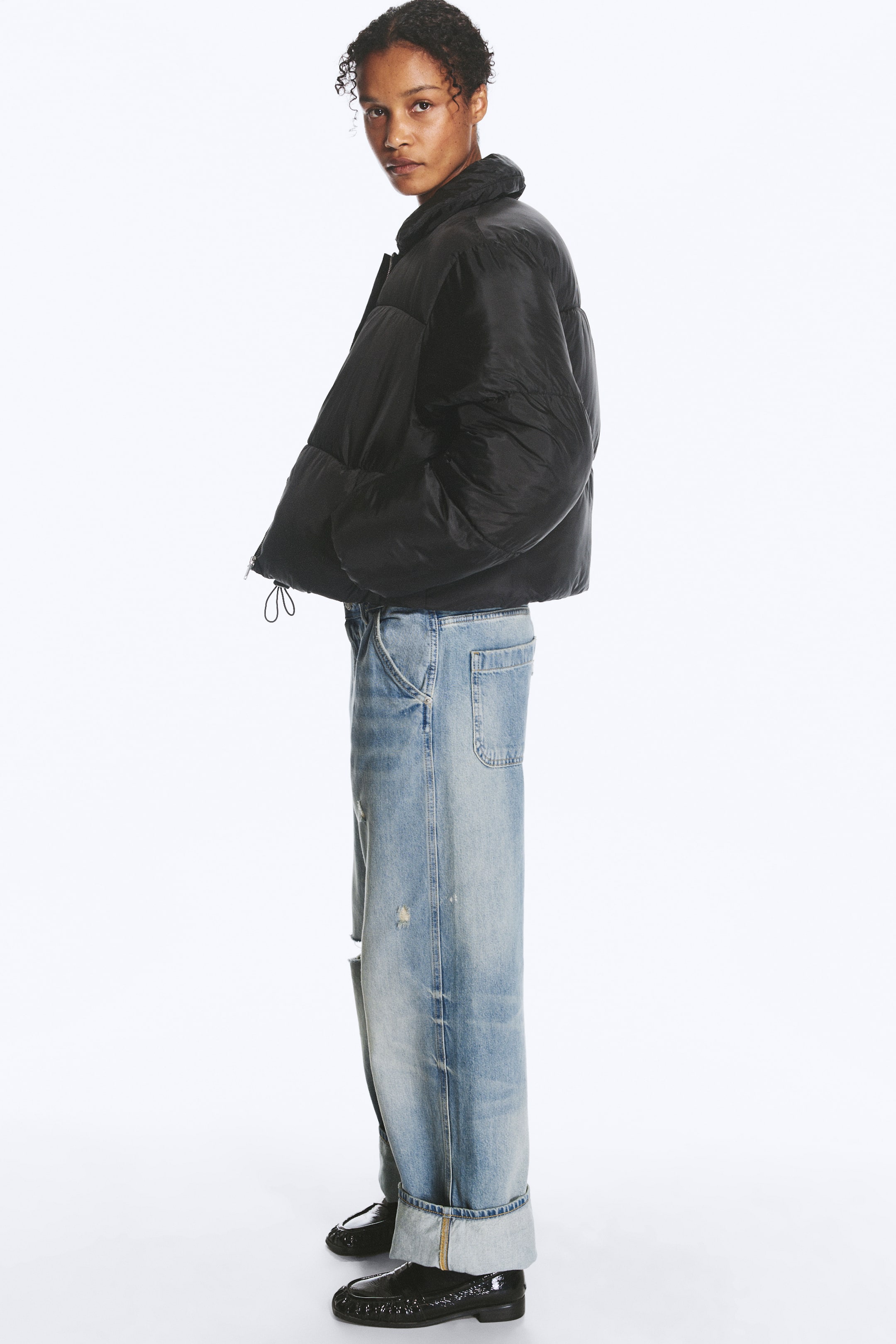 Größeres Bild ansehen: Eine schwarze Steppjacke mit wattiertem Kragen, einem Reißverschluss vorne und einem sichtbaren elastischen Kordelzug in der Taille wird vom Model getragen. Das Model steht im Profil, die Hände in den Seitentaschen, und kombiniert die Jacke mit hell gewaschenen, weit geschnittenen Jeans mit Umschlag und schwarzen Schuhen.