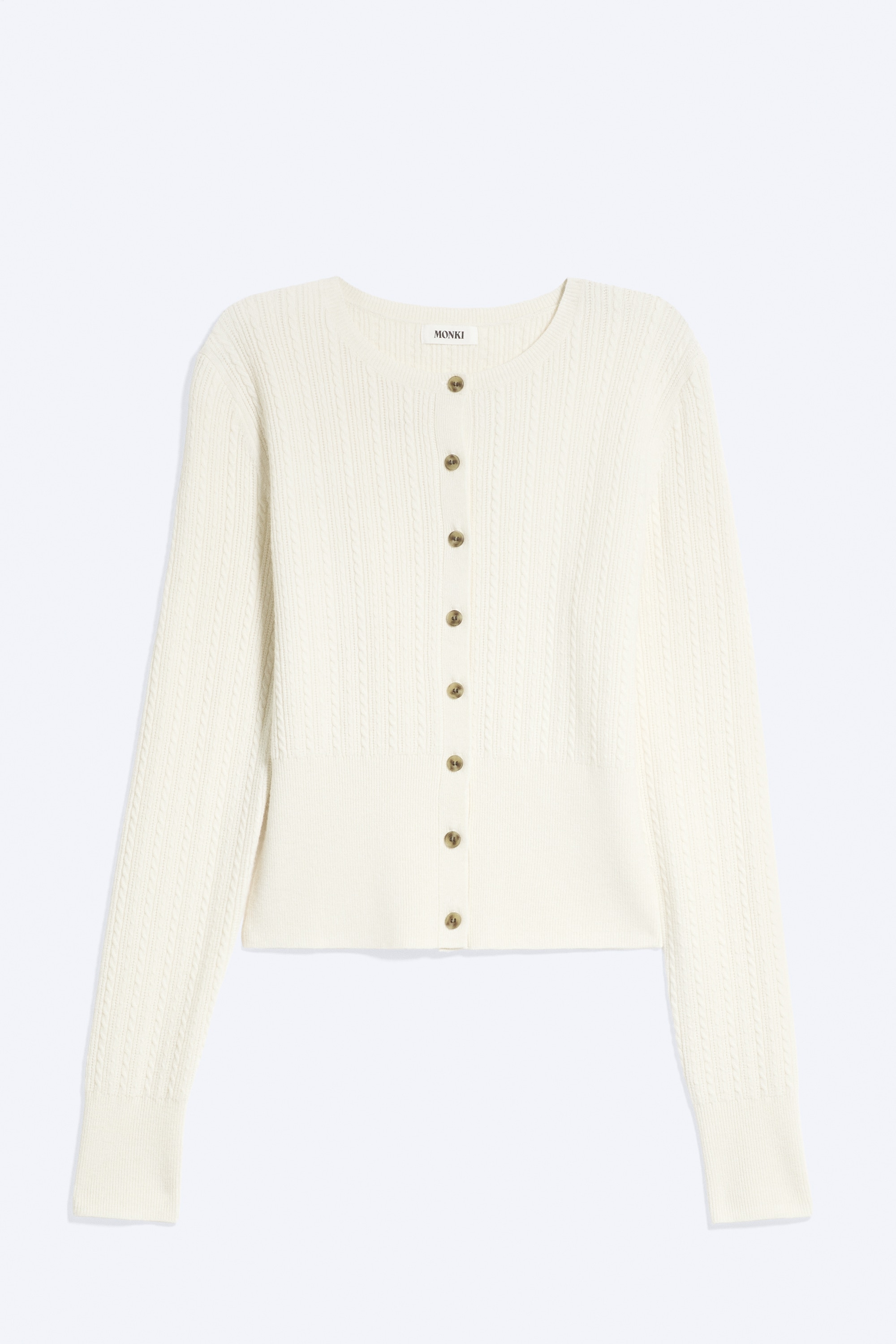 Un cardigan crema lavorato a trecce con bottoni marroni è indossato dalla modella, che tiene con entrambe le mani l’abbottonatura frontale. Il cardigan slim fit presenta uno scollo tondo e maniche lunghe con polsini a coste ampi, ed è abbinato a pantaloni in maglia color crema coordinati.