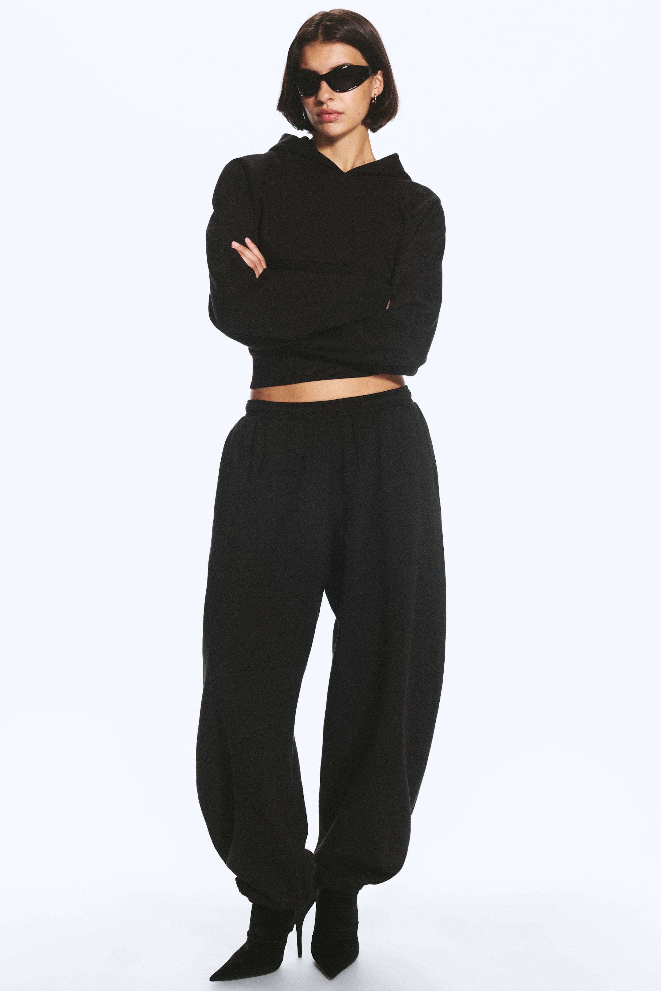 Ver imagem maior: Calças de fato de treino pretas, de corte folgado, com cintura elástica e punhos franzidos nos tornozelos, usadas pelo modelo. As calças de fato de treino estão combinadas com um hoodie preto curto a condizer e botins pretos de salto com biqueira bicuda.