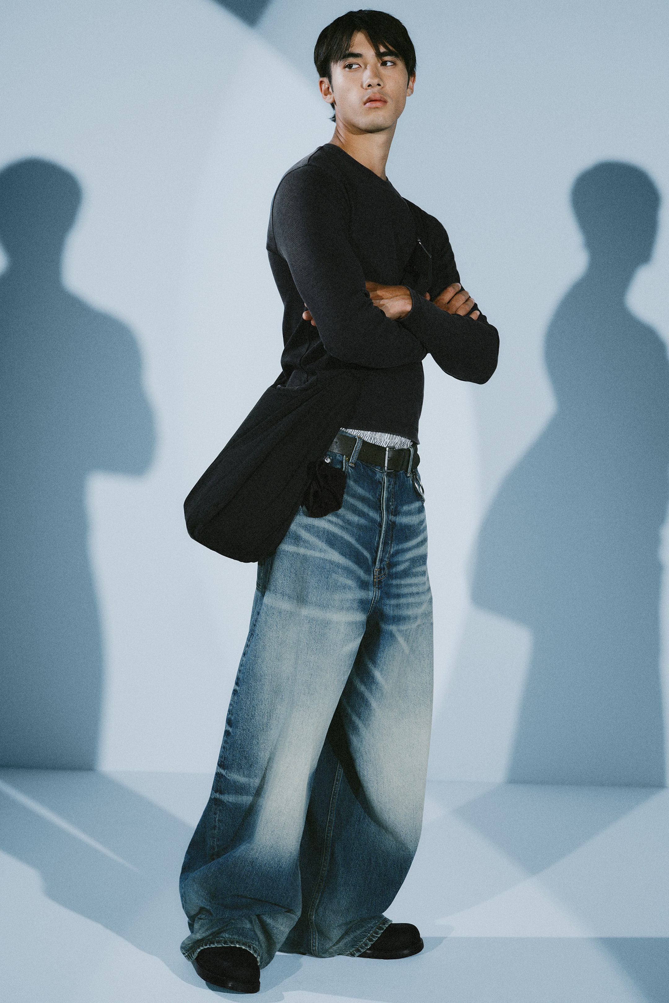Grotere afbeelding bekijken: Een paar blauwe low-rise jeans is weergegeven vanuit een frontale driekwarthoek, met een baggy barrel-leg silhouet met duidelijke fading en whiskering op de dijen. De jeans worden gedragen met een zwarte leren riem, die de low-rise tailleband benadrukt, en vallen losjes over zwarte schoenen.