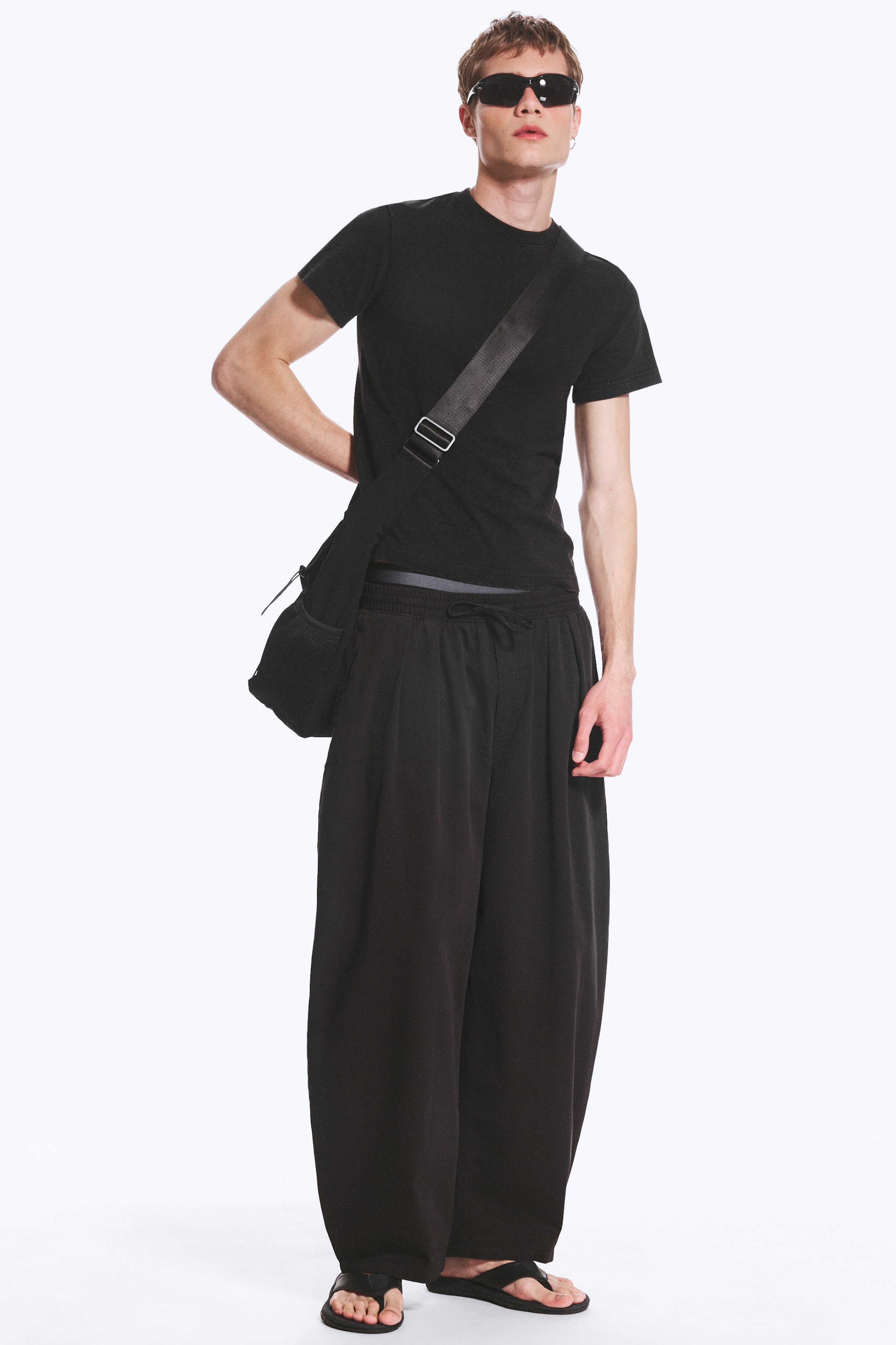 Grotere afbeelding bekijken: Zwarte wijde broek wordt gedragen door het model, met een ontspannen pasvorm, een trekkoord in de taille en een volledige lengte. Een zwarte crossbody tas met een brede band wordt over het lichaam van het model gedragen, rustend op de heup.