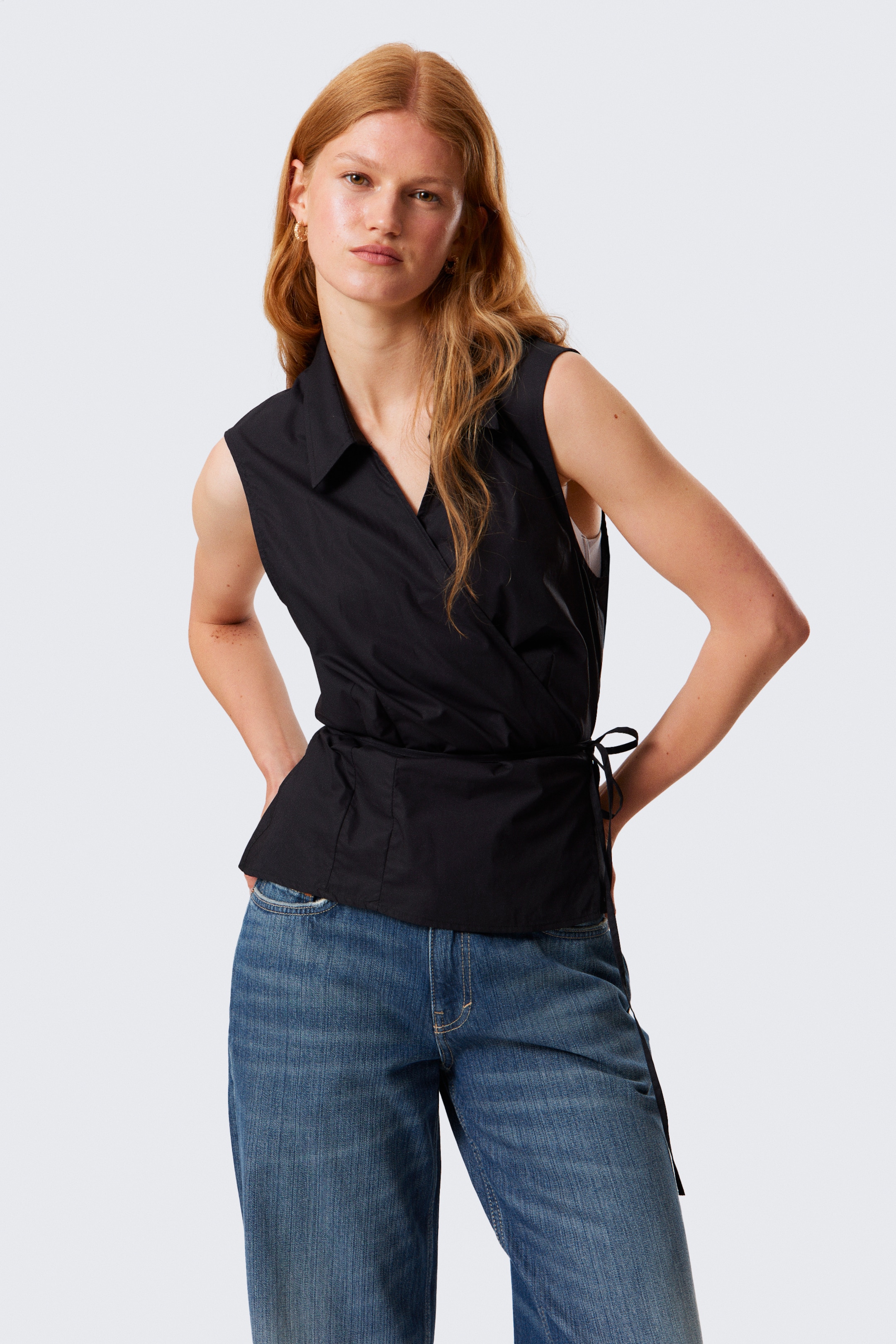 Ingrandisci l'immagine: Un top nero senza maniche avvolgente, realizzato in cotone leggero e tagliato per una vestibilità aderente, è indossato dalla modella. Il top presenta un colletto a punta, un design avvolgente con lacci laterali regolabili e un orlo pulito, e la modella è in piedi con le mani nelle tasche dei jeans blu.