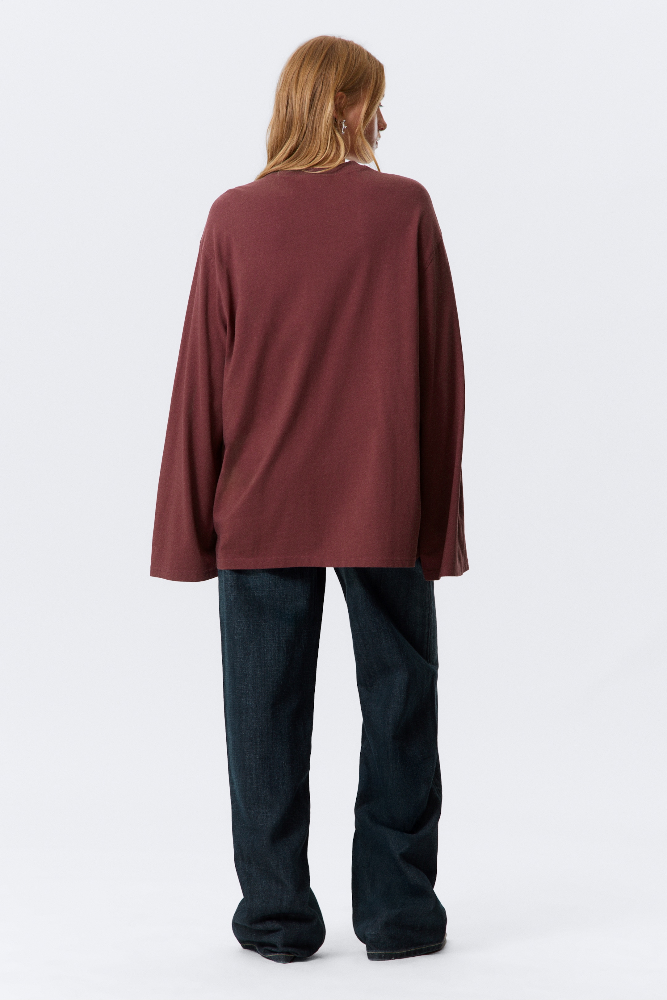 Visualizza immagine più grande: Il retro di una maglia oversize a maniche lunghe di un marrone rossastro intenso è visibile, tagliata con una vestibilità ampia e maniche larghe che cadono sulle spalle. L'orlo dritto della maglia cade sui fianchi ed è indossata sopra jeans scuri a gamba larga.