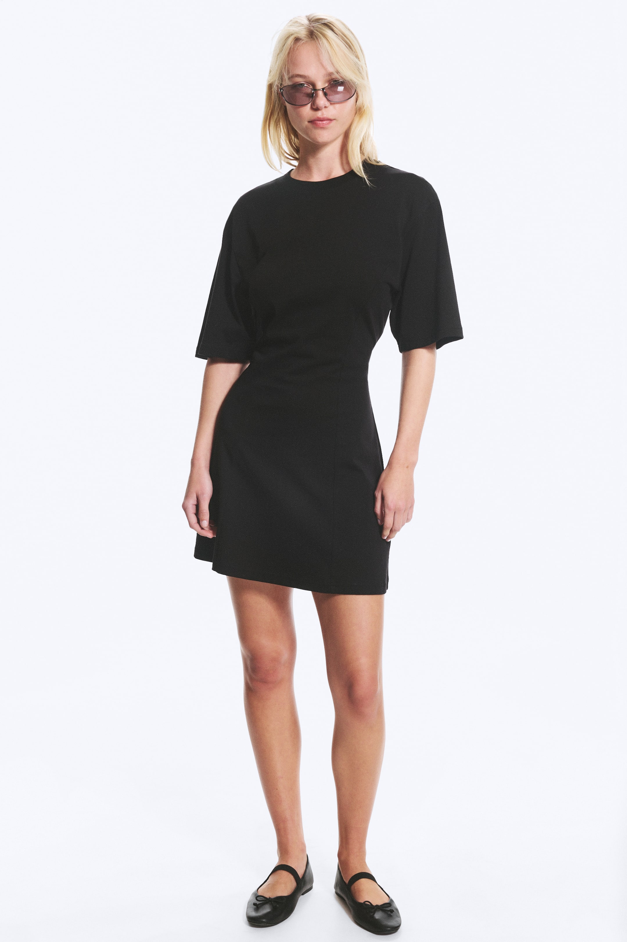 Ver imagen más grande: Un vestido negro mini de corte fit-and-flare tipo camiseta, con cuello redondo, mangas cortas voluminosas, cintura ajustada y falda acampanada, está siendo lucido por la modelo.