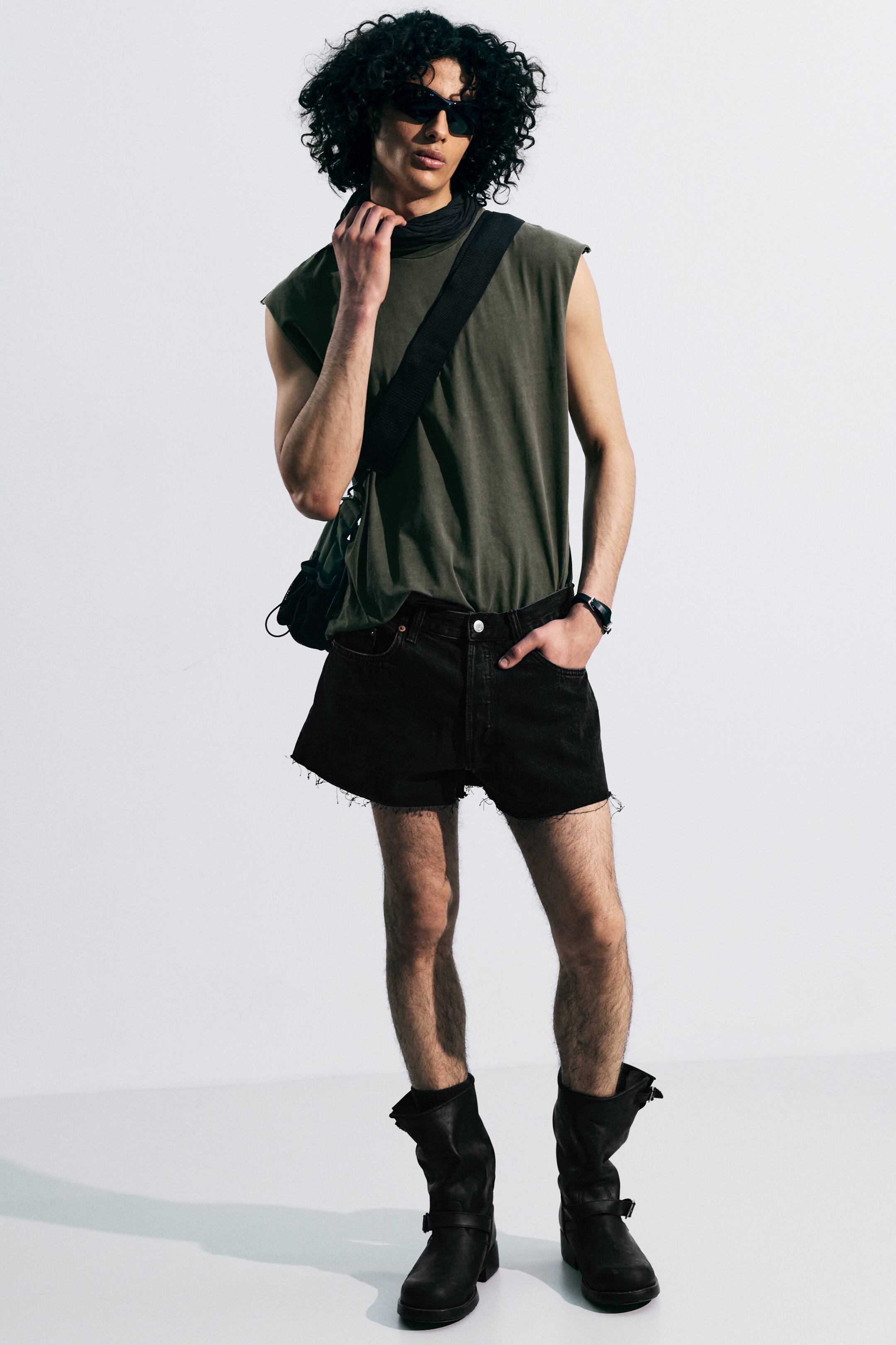 Agrandir l'image: Un haut sans manches vert olive, avec une coupe décontractée et un col montant noir en dessous, est rentré dans un short en jean noir taille haute. Le short présente une coupe ample et un ourlet brut et effiloché, tandis que des bottes noires mi-mollet, avec des sangles à boucle et des tiges souples, complètent l'ensemble.