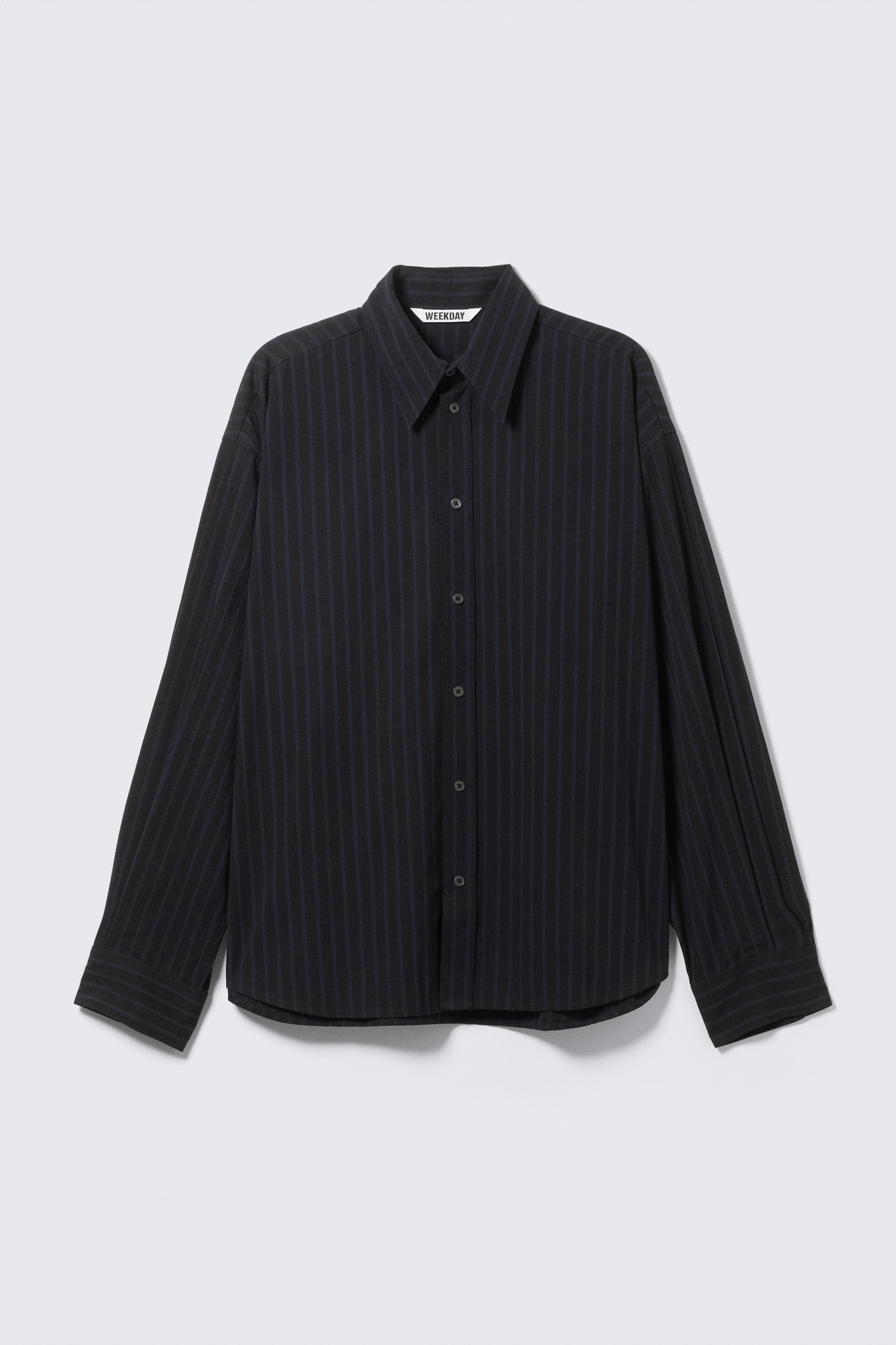 Ver imagen más grande: Una camisa negra de manga larga muestra su parte delantera, con sutiles rayas verticales. Está diseñada con un cuello en punta, un cierre frontal de botones completos y mangas largas con puños abotonados. Una etiqueta de la marca "WEEKDAY" es visible dentro del cuello, y el dobladillo es curvo.