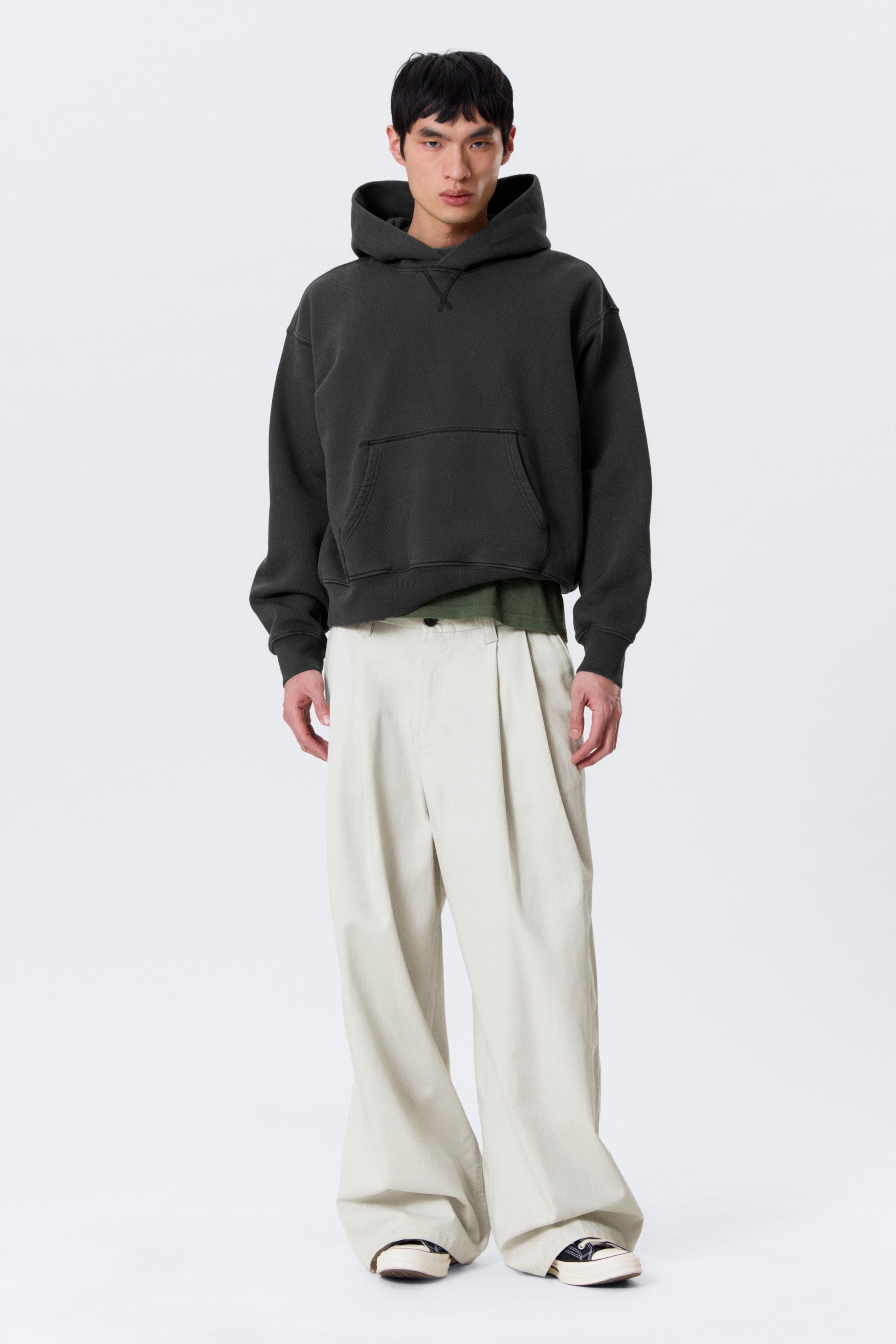 Visa större bild: En mörkgrå huvtröja presenteras framifrån, med en känguruficka, ribbade muddar och fåll, samt en V-söm detalj under halsringningen. Den är lagrad över en grön t-shirt och kombinerad med ljusbeige, vida veckade byxor som vilar över skorna.