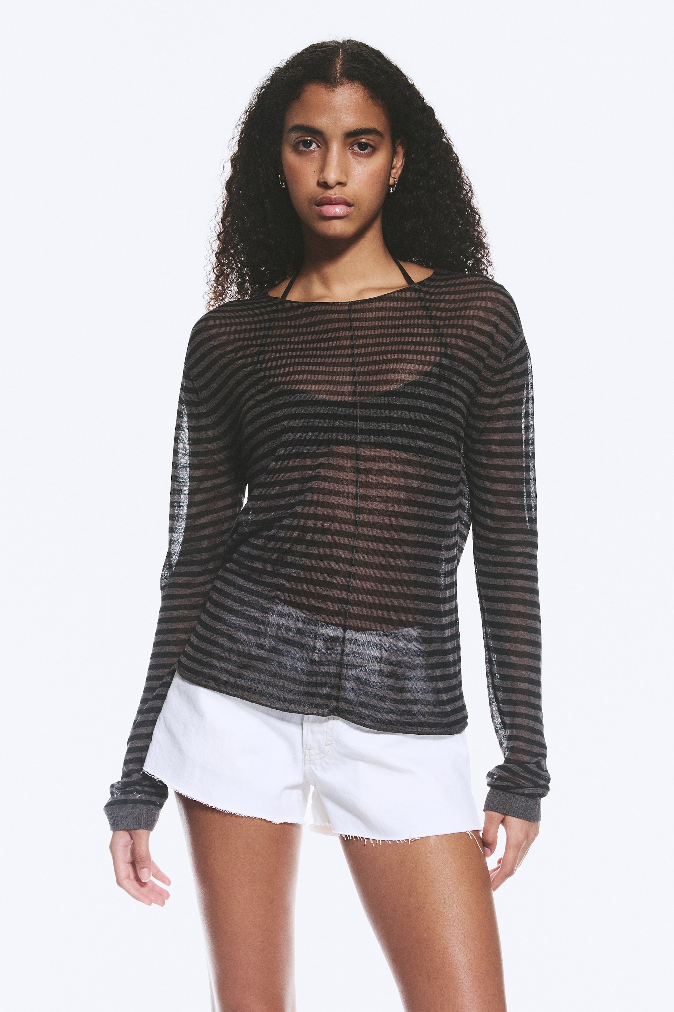 Zobraziť väčší obrázok: Modelka má na sebe voľne padnúci, priehľadný, pruhovaný pletený sveter so širokým výstrihom, spustenými ramenami a rebrovanými manžetami. Pod priehľadným materiálom je vidieť tmavú podprsenku a biele šortky dopĺňajú outfit modelky.