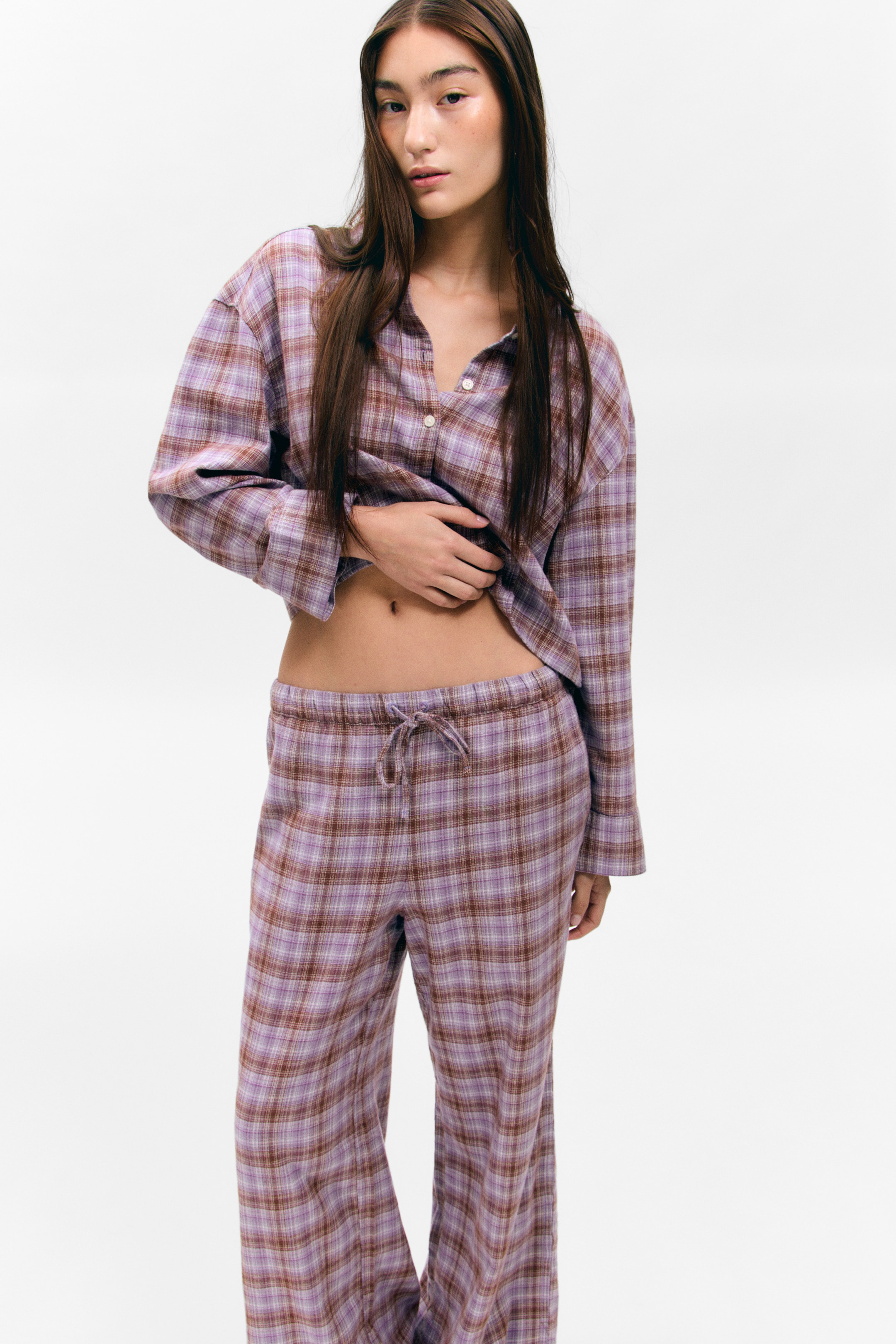 View larger image: A purple and brown plaid pajama set is being presented from a front-facing angle. The relaxed-fit, long-sleeved button-up shirt is featuring white buttons and is being lifted at the hem, while the matching pants are displaying a drawstring elastic waistband.