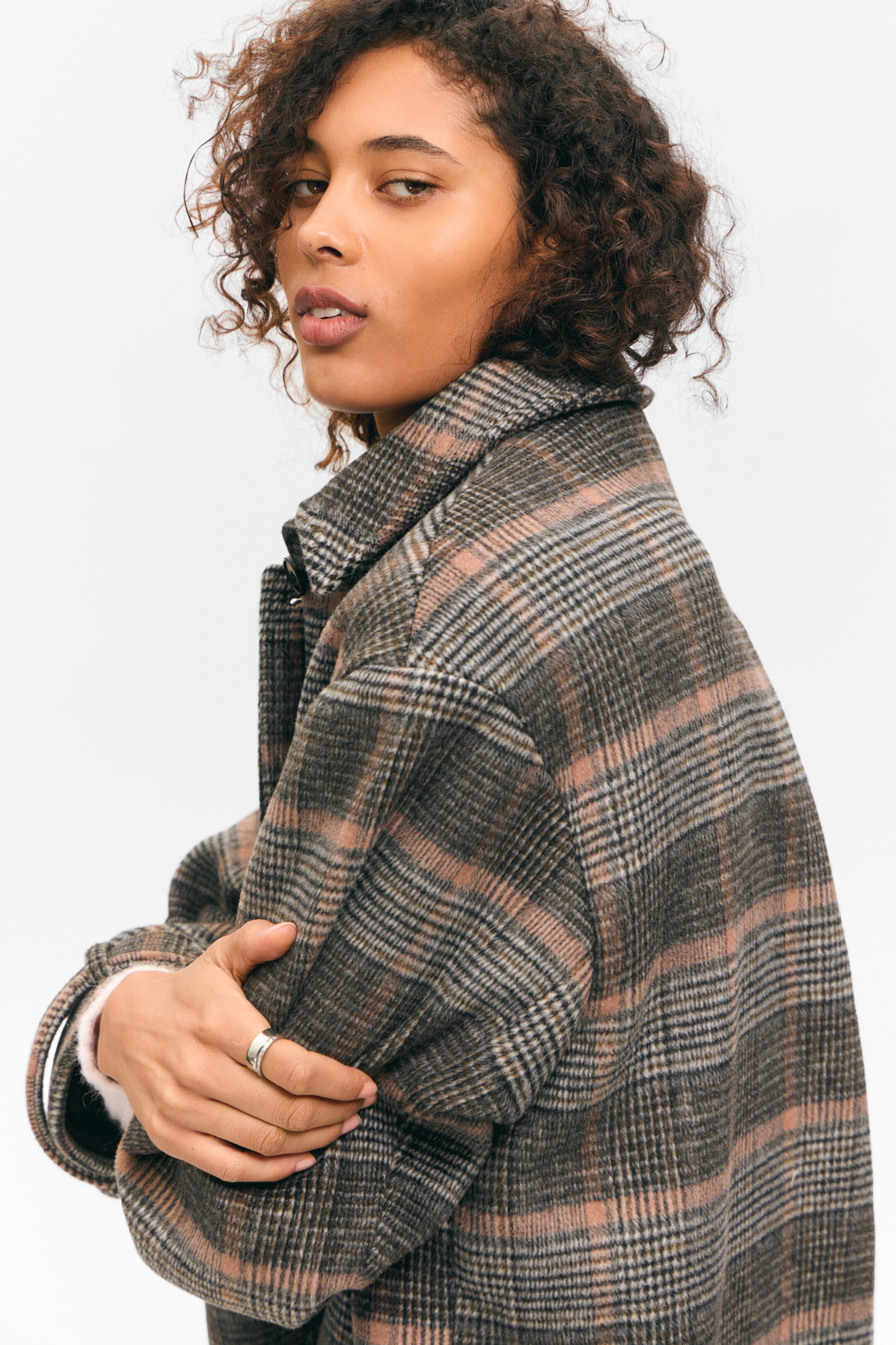 Größeres Bild ansehen: Eine karierte Jacke mit Kragen wird modelliert, von hinten links gesehen, mit einem strukturierten Stoff in Braun, Schwarz, Beige und Hellrosa Karo. Die Jacke hat eine entspannte Passform, und ein helles, langärmeliges Kleidungsstück wird unter ihrer Manschette geschichtet.