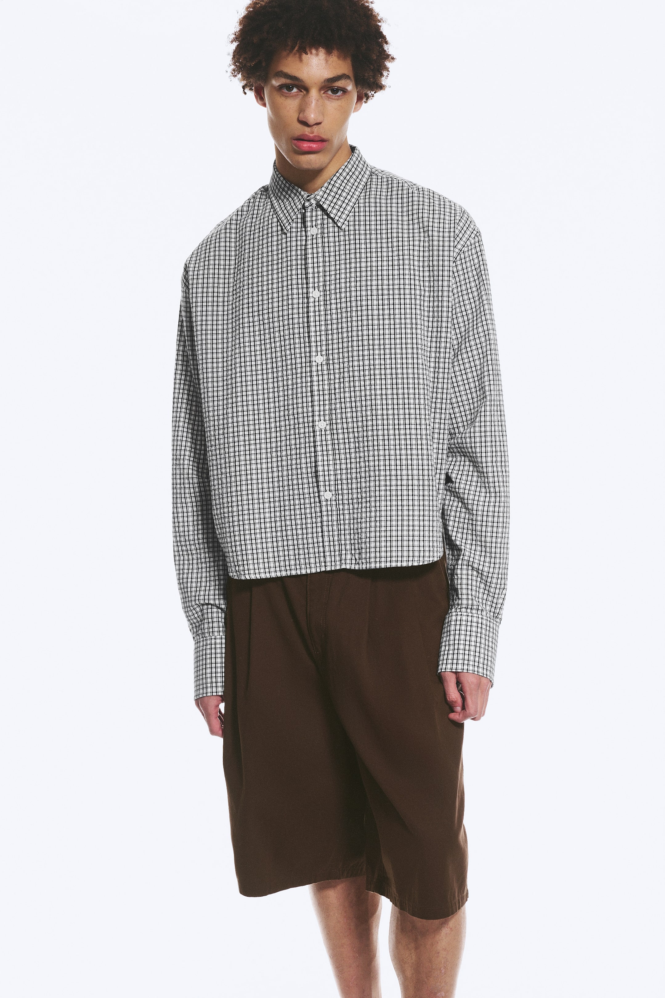 Agrandir l'image: Une chemise blanche avec un petit motif à carreaux noirs présente un col standard, une fermeture à boutons sur le devant, des manches longues avec poignets boutonnés et un ourlet court. Elle est portée avec un short plissé marron foncé atteignant le genou.