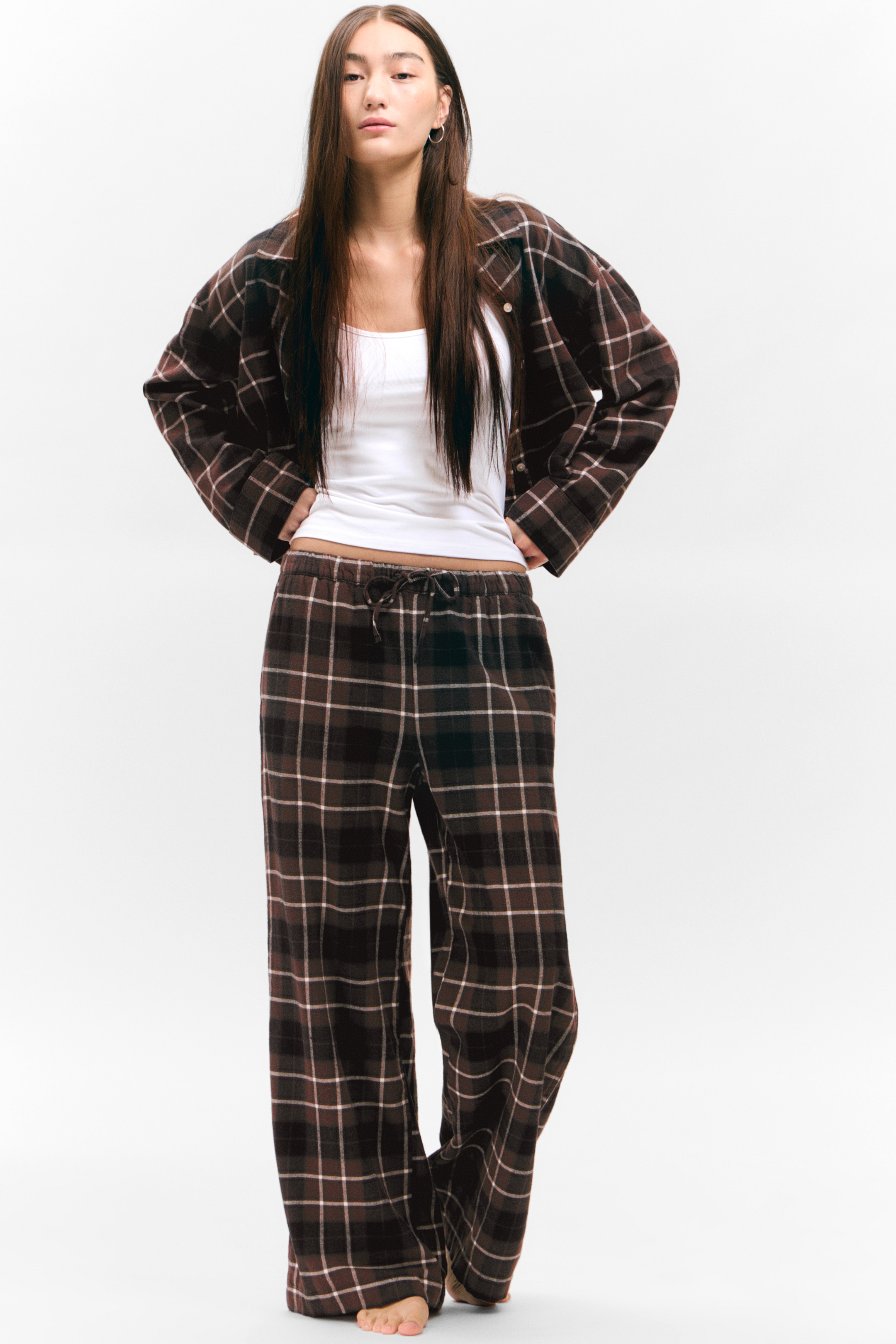 Größeres Bild ansehen: Ein dunkelbraunes, schwarzes und cremefarbenes kariertes Pyjama-Set wird von vorne präsentiert. Das Hemd mit Kragen wird offen über einem weißen Tanktop getragen, und die dazu passenden weit geschnittenen Hosen verfügen über einen Kordelzug mit elastischem Bund.