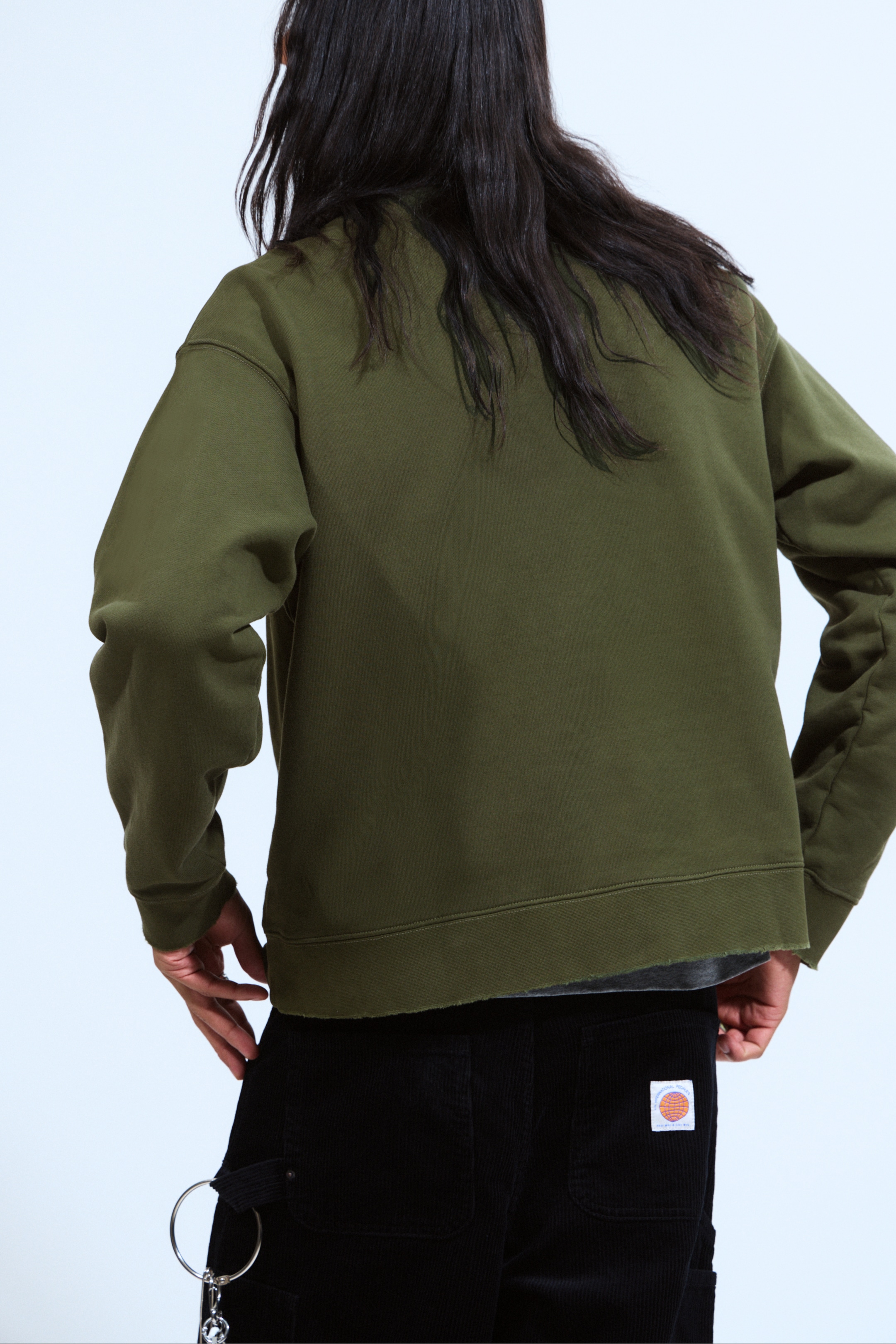 Agrandir l'image: Un sweat-shirt vert olive foncé est présenté de dos, avec une coupe décontractée et des manches longues avec des poignets côtelés. Son ourlet à bord brut repose à la taille, laissant apparaître un t-shirt gris en dessous.