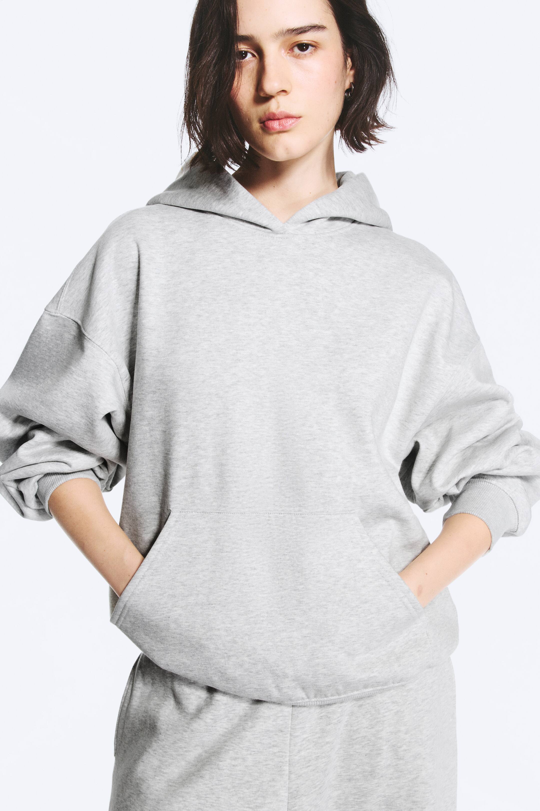 Преглед на по-голямо изображение: Моделът е облечен в oversized суитшърт с качулка в светлосив цвят и е застанал с двете ръце, поставени във предния кенгуру джоб. Суитшъртът с качулка е с паднали рамене, свободна кройка и качулка без връзки, и е съчетан с подходящи светлосиви спортни панталони.