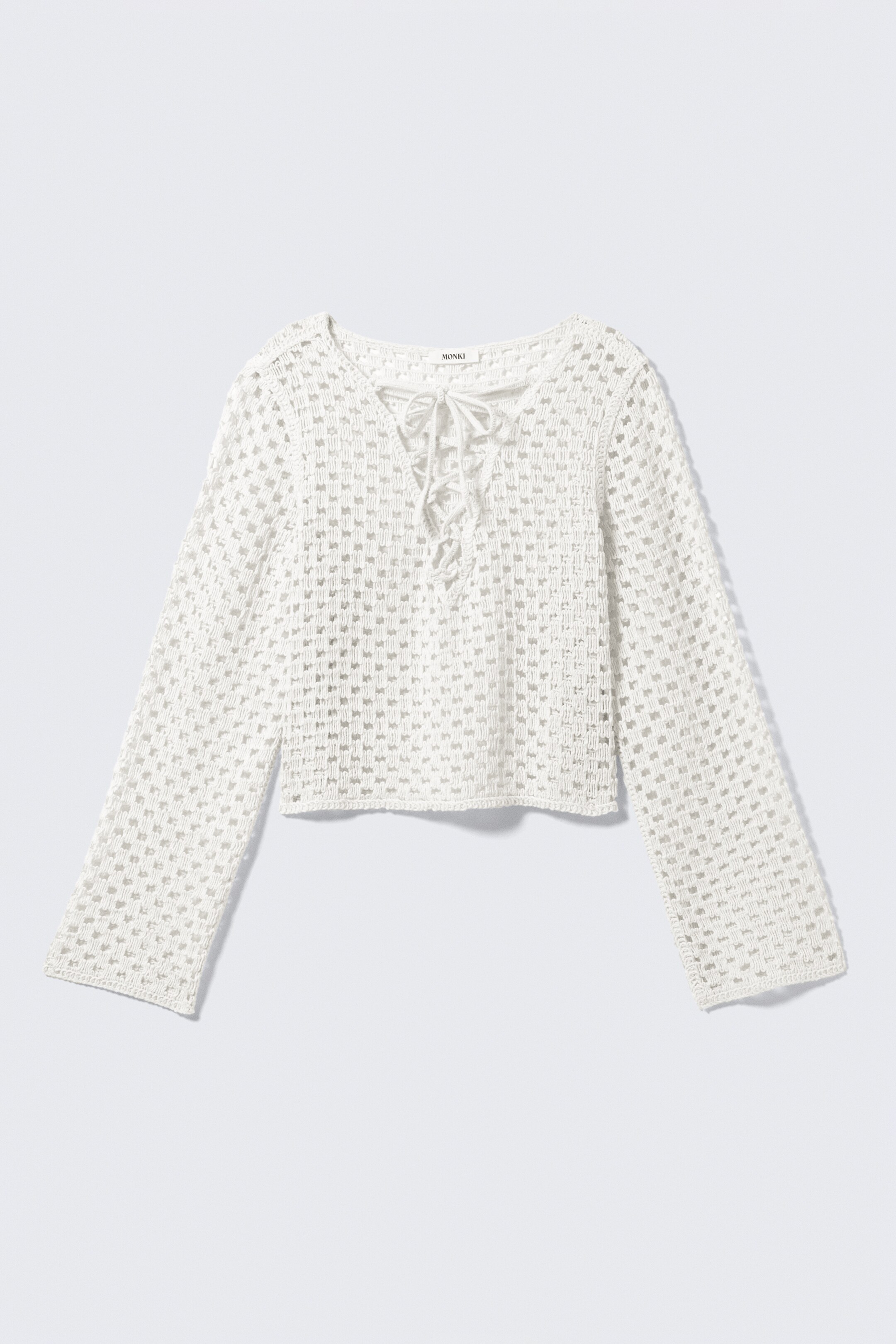 Ver imagem maior: Um top branco de mangas compridas, corte descontraído e ligeiramente curto está representado pela FRENTE, apresentando um tecido de malha com aparência de crochet com detalhes em aberturas, um decote em V com frente entrançada e laços finos, e um rebordo de gola elegante.